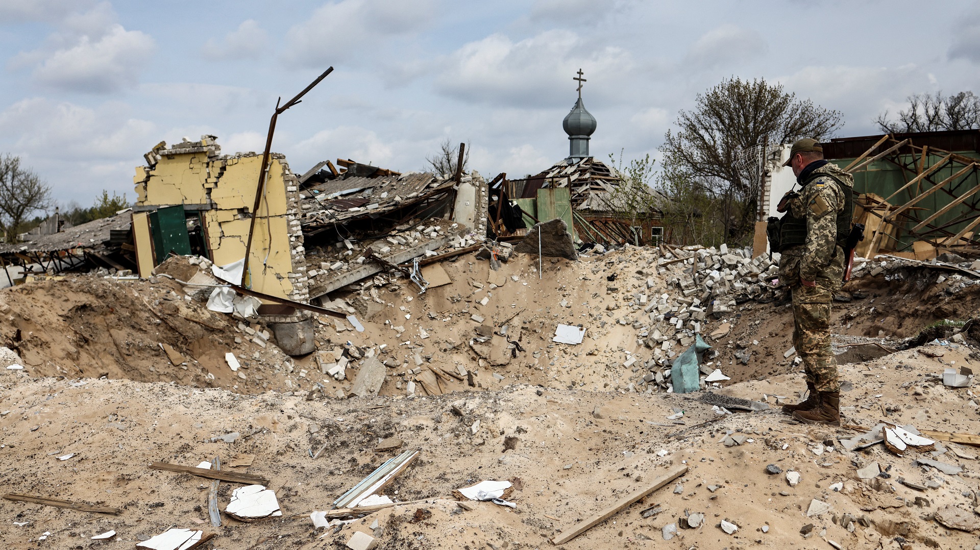 A Ukranian serviceman looks into a crater and a destroyed home are pictured in the village of Yatskivka, eastern Ukraine on April 16, 2022. - Russia's military focus now seems to be on seizing the eastern Donbas region, where Russian-backed separatists control the Donetsk and Lugansk areas. Lugansk governor Serhiy Gaidai called on April 16, 2022, for civilians to leave the area while they still can. (Photo by RONALDO SCHEMIDT / AFP)