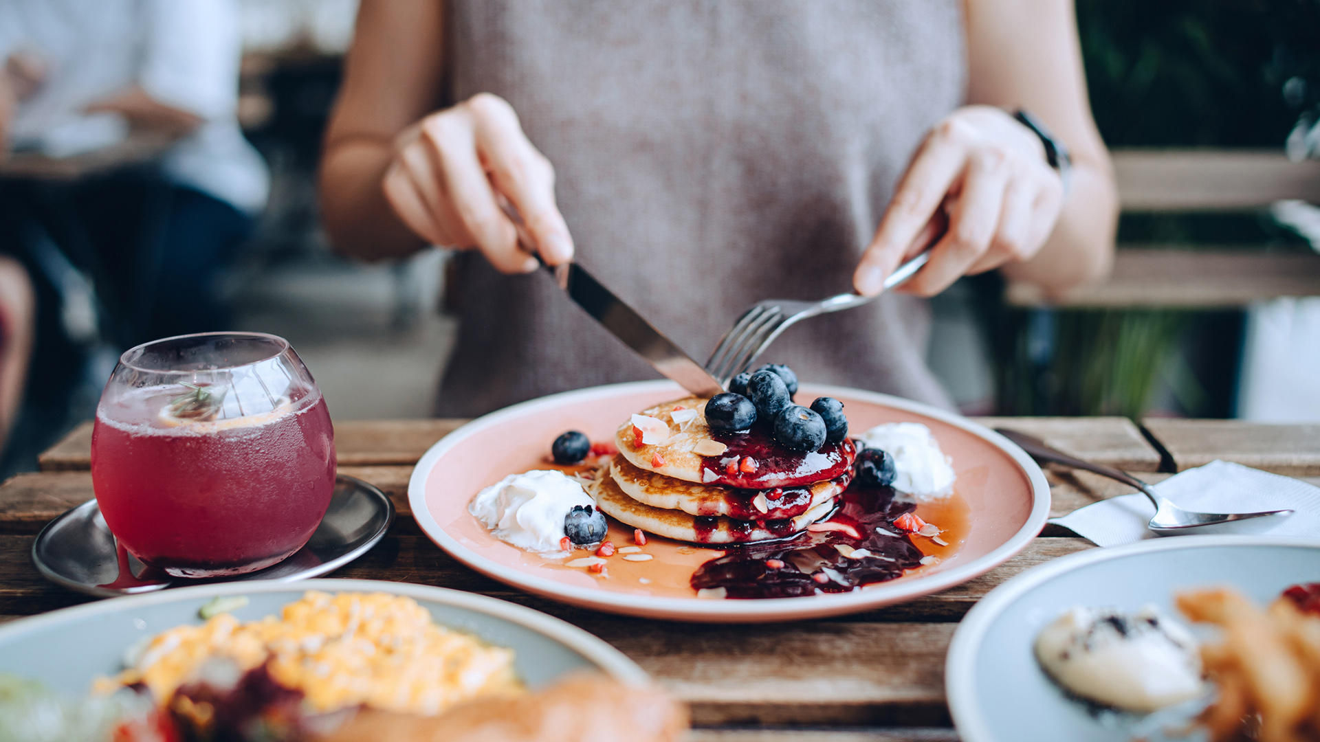 Close up of young woman sitting at dining table eating pancakes with blueberries and whipped cream in cafe, with English breakfast and french fries served on the dining table. Eating out lifestyle