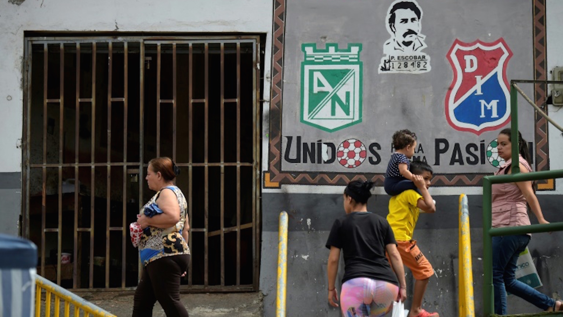 Vecinos del barrio Pablo Escobar pasan por delante de una pintura con la cara del capo del narcotráfico, junto a los escudos del DIM y de Atlético Nacional (AFP - Raul ARBOLEDA)  