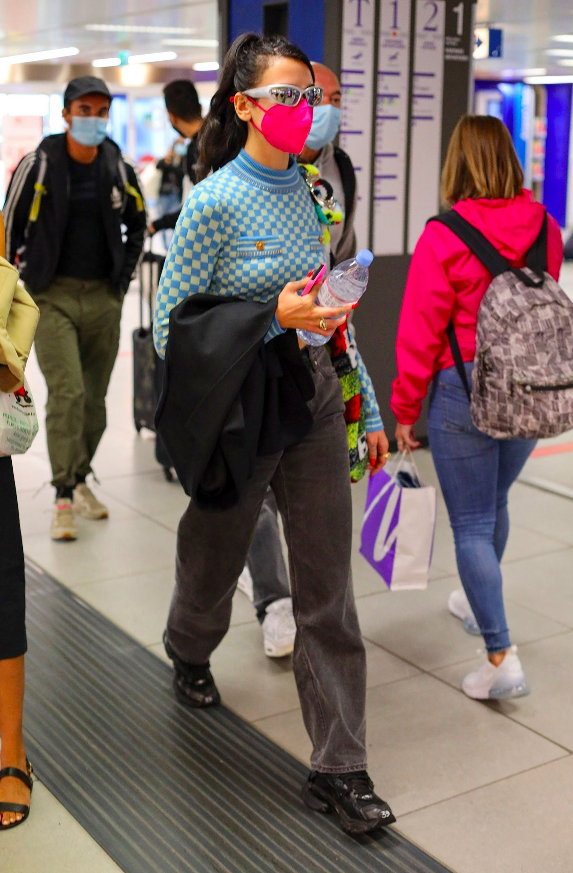 Dua Lipa fue fotografiada en el aeropuerto luego de su paso por La Semana de la Moda en Milán. La cantante no pasó desapercibida con su look: lució un jean negro, remera con rombos celestes y botones dorados, cartera colorida y tapabocas fucsia