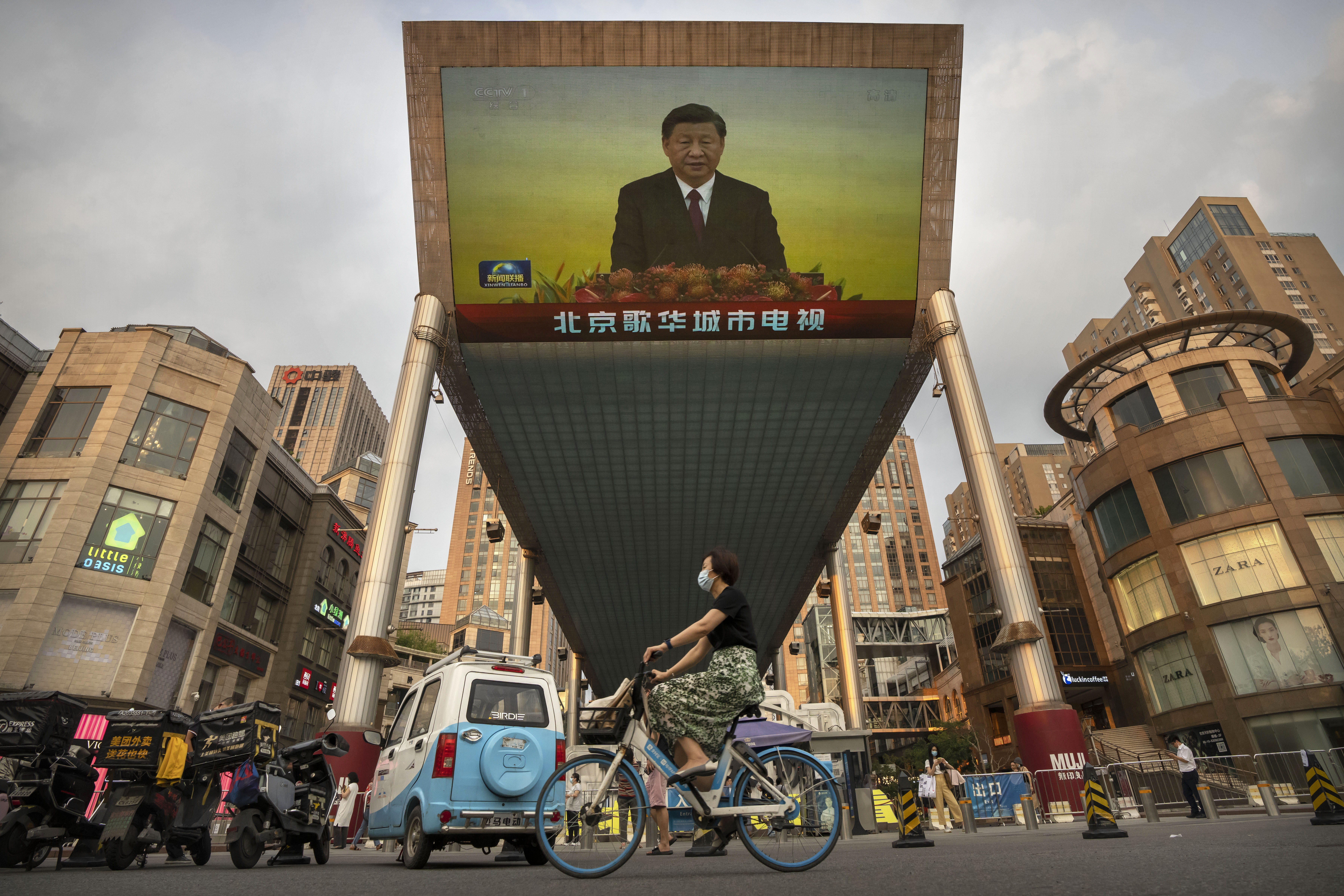 Una mujer con mascarilla circula en bicicleta bajo una gran pantalla de televisión en Pekín que muestra las noticias de la televisión estatal china sobre la visita del presidente Xi Jinping a Hong Kong el 1 de julio de 2022 (AP Foto/Mark Schiefelbein)