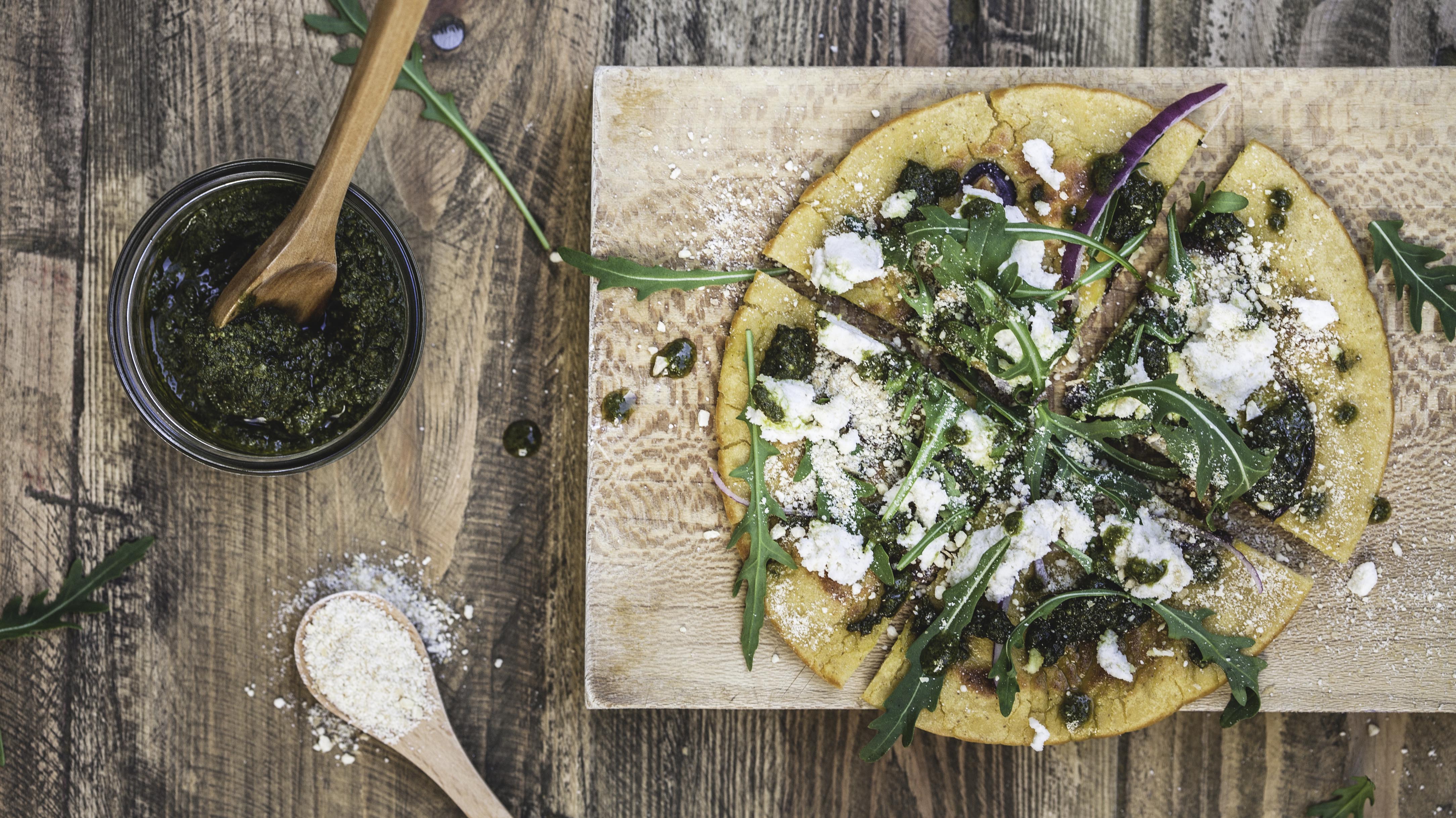 A chickpea flour based pizza, fried in a cast-iron skillet with red onions in the base and finely shredded on the top. A pesto made from carrot tops and vegan parmesan cheese is dabbed on top with smears of almond-based ricotta cheese and handfuls of wild arugula/rocket leaves.