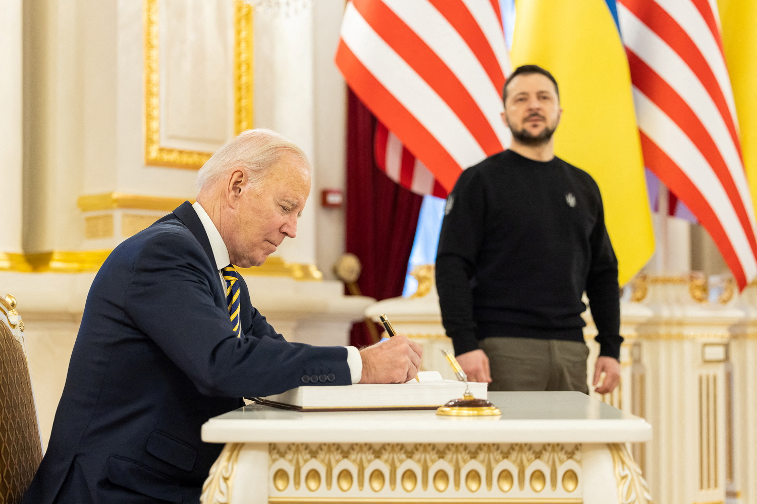El presidente de los Estados Unidos deja un mensaje en un libro mientras el presidente de Ucrania se encuentra a su lado.
