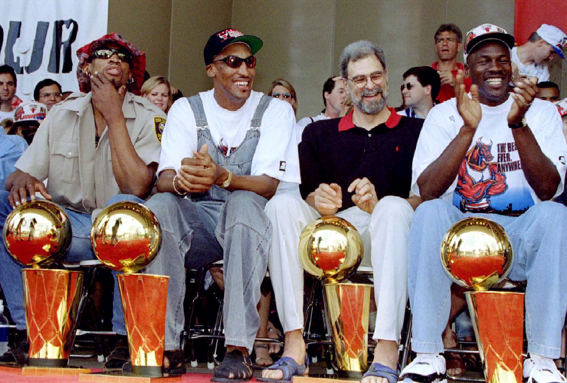 Con sus cuatro trofeos de campeonato a sus pies, los Chicago Bulls Dennis Rodman (izquierda), Scottie Pippen (izquierda, centro), el entrenador Phil Jackson y Michael Jordan (derecha) se ríen de los comentarios de un compañero de equipo en un mitin del equipo en Chicago.