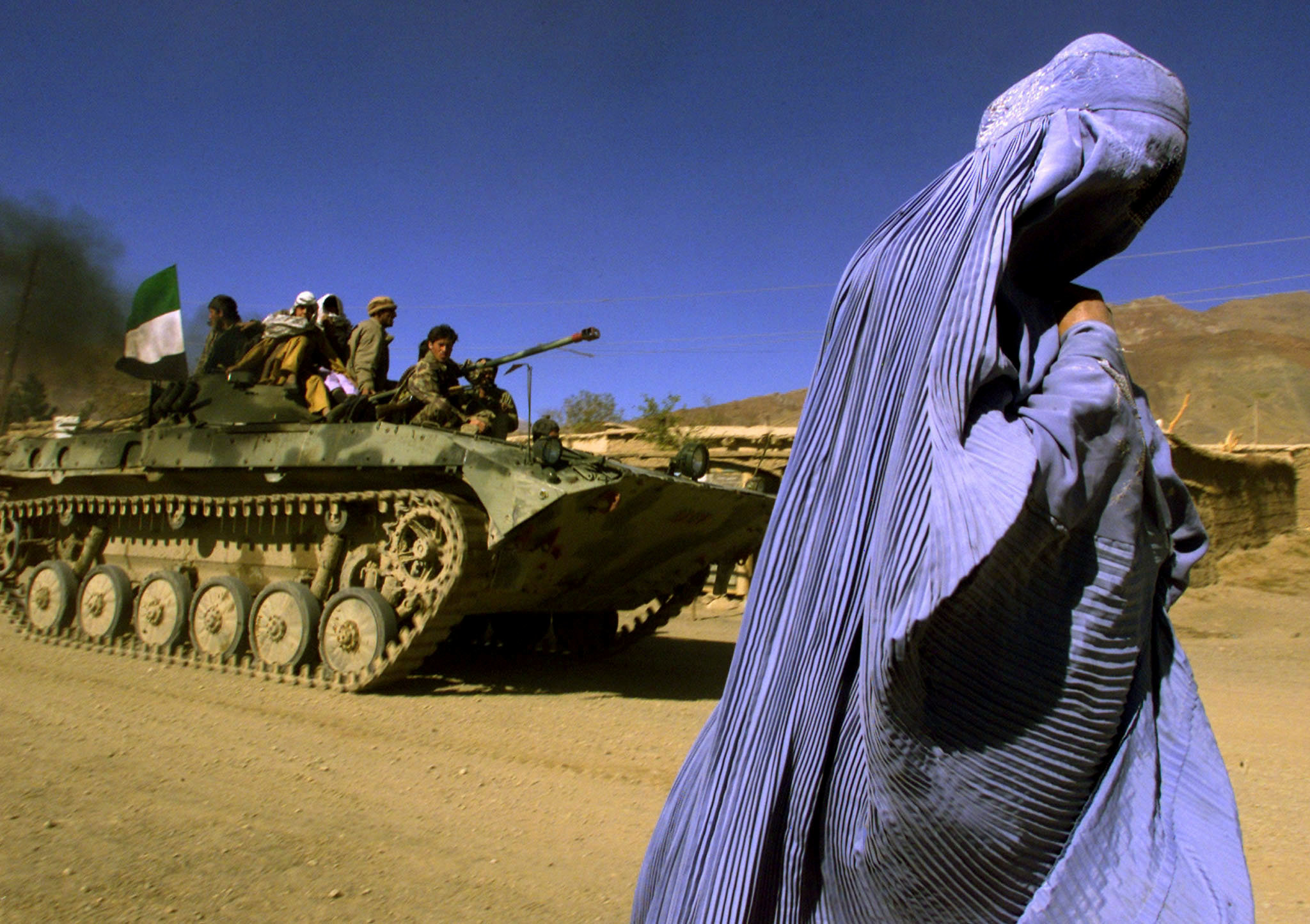 Una mujer afgana vestida con un burka tradicional camina al costado de una carretera mientras un APC (transporte blindado de personal) de la Alianza del Norte, que lleva combatientes y la bandera afgana, se dirige a una nueva posición en las afueras de Jabal us Seraj, a unos 60 km. al norte de la capital afgana, Kabul, 4 de noviembre de 2001. (REUTERS / Yannis Behrakis/archivo)