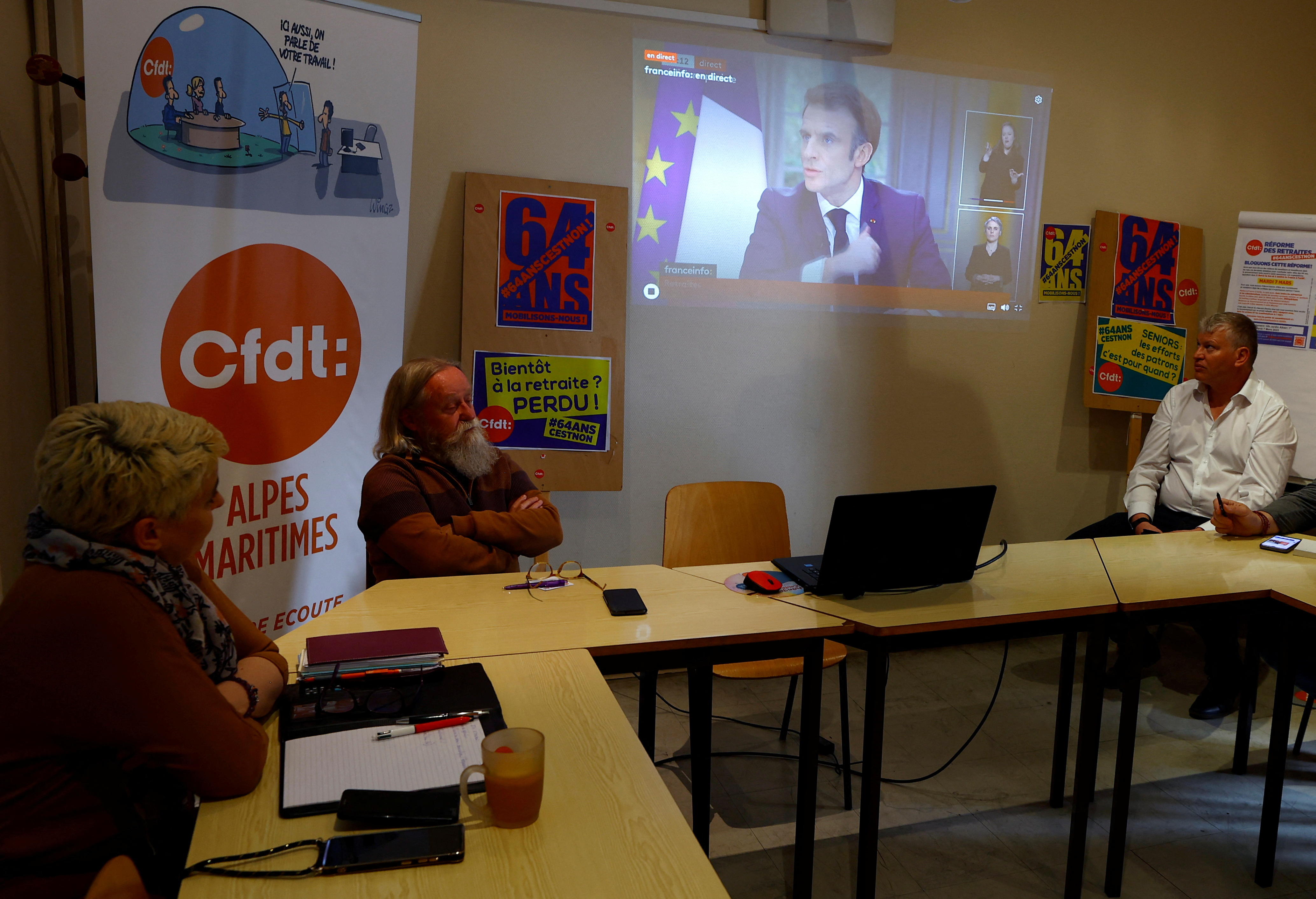 Miembros del sindicato CFDT observan al presidente francés Emmanuel Macron en una pantalla mientras habla durante una entrevista en la televisión nacional para esbozar los planes tras la aprobación de la reforma de las pensiones al rechazar el Parlamento francés dos mociones de censura contra el Gobierno, en Niza, Francia, 22 de marzo de 2023. REUTERS/Eric Gaillard