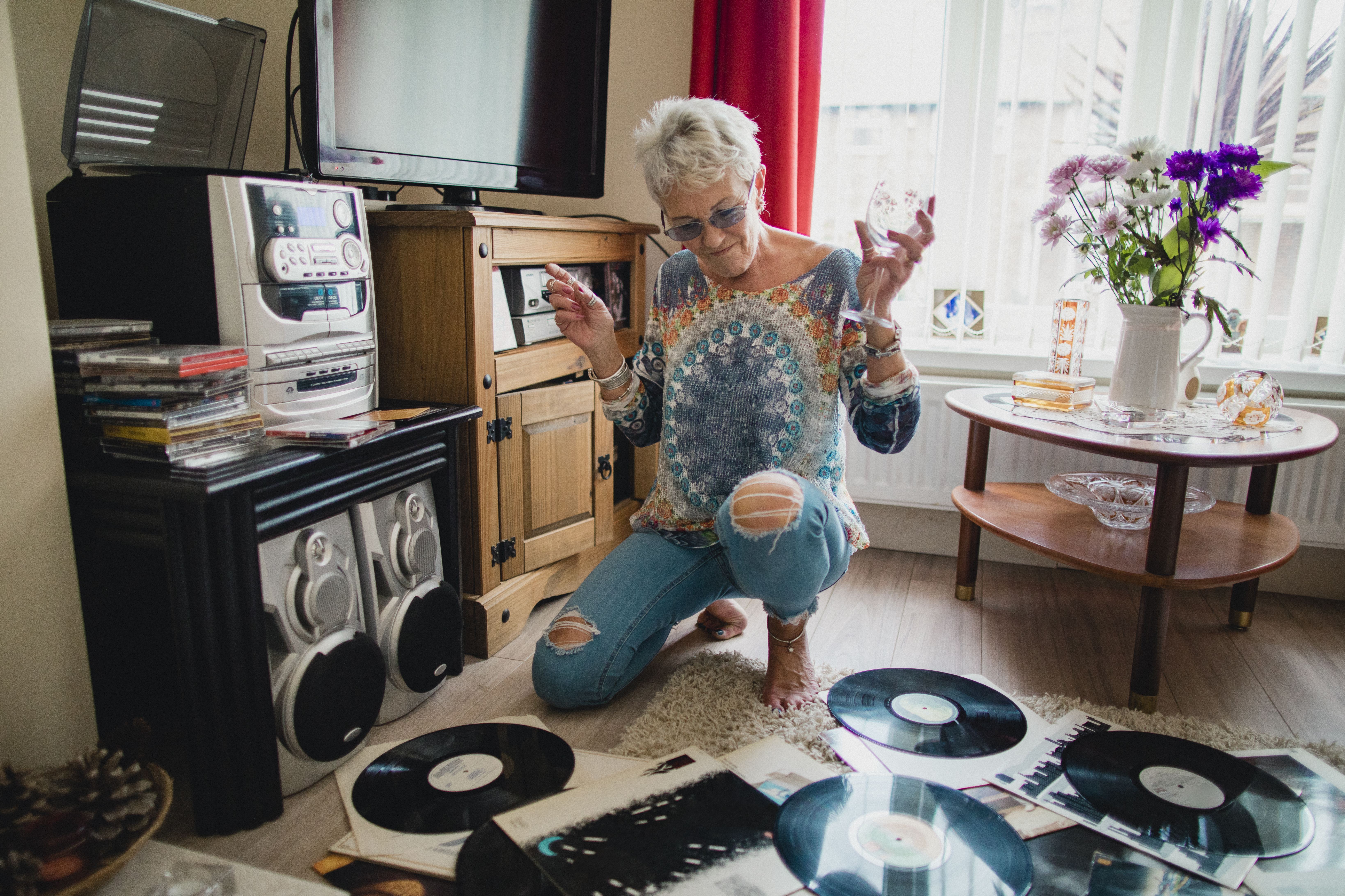 One senior woman sitting on the floor in her living room, she is surrounded by vinyl records and is listening to music from a record player.