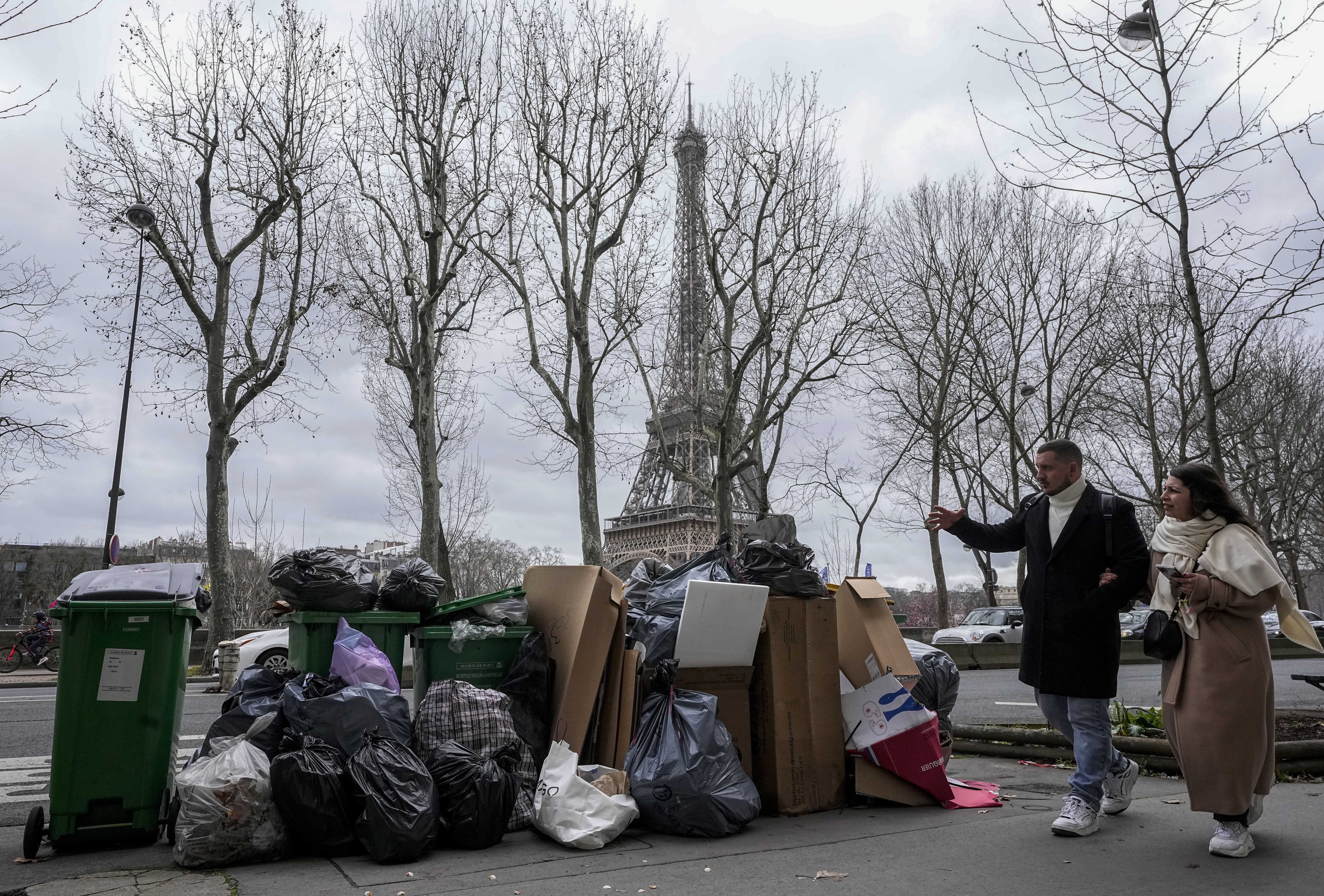 Two people walk past overflowing trash cans near the Eiffel Tower in Paris on March 12, 2023. (AP Photo/Michel Euler)