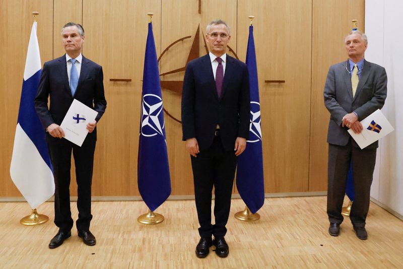 Los embajadores de Finlandia (izquierda) y Suecia (derecha) ante la OTAN flanquean al secretario general de la OTAN, Jens Stoltenberg, mientras sostienen sendas carpetas con sus respectivas banderas nacionales en la ceremonia de entrega de la solicitud de adhesión de ambos países a la alianza militar, celebrada en Bruselas, Bélgica, el 18 de mayo de 2022. 
