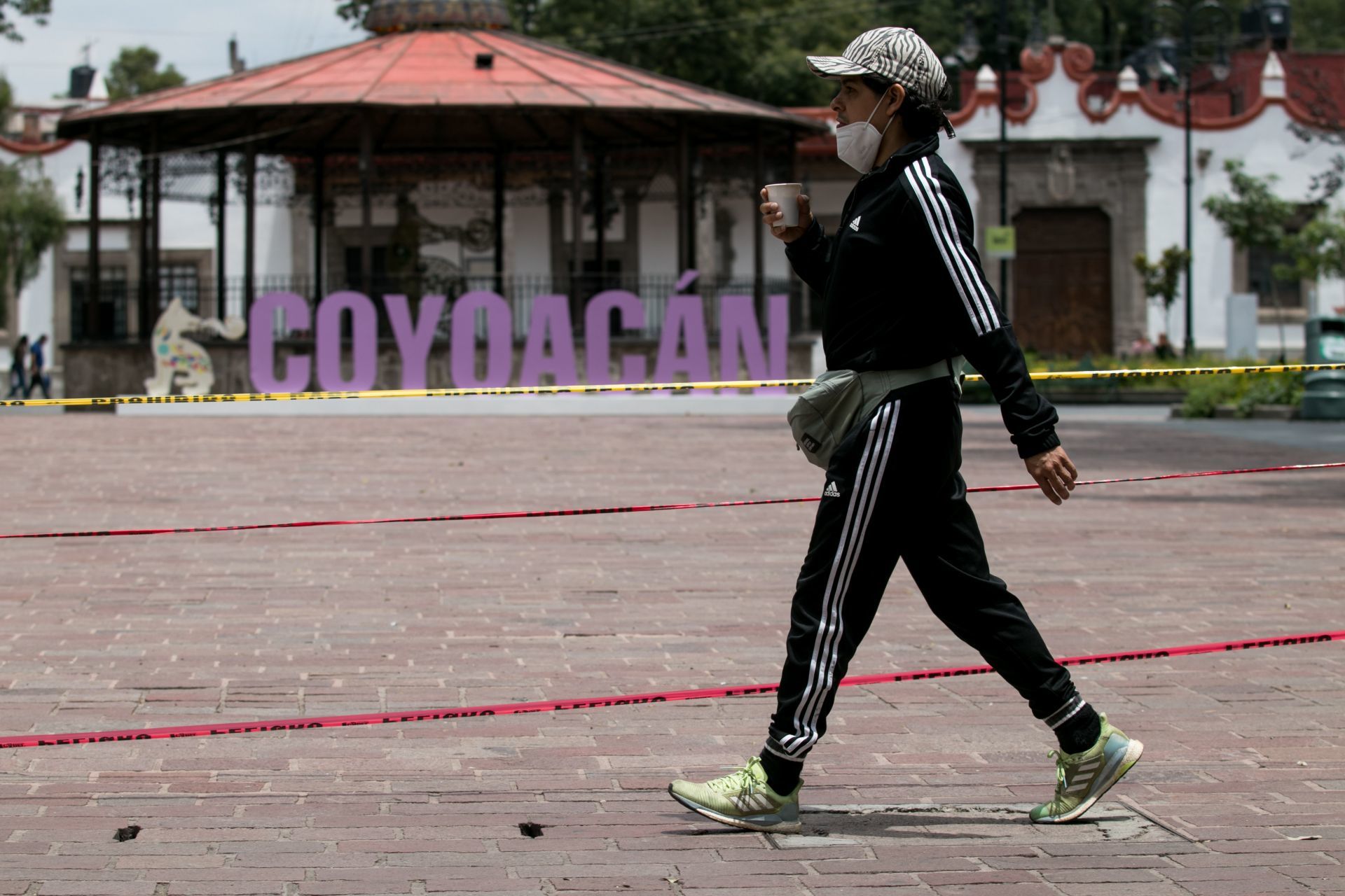 Un hombre con su cubrebocas mal colocado bebe café mientras recorre el centro de Coyoacán, el cual se encuentra acordonado para evitar aglomeraciones.