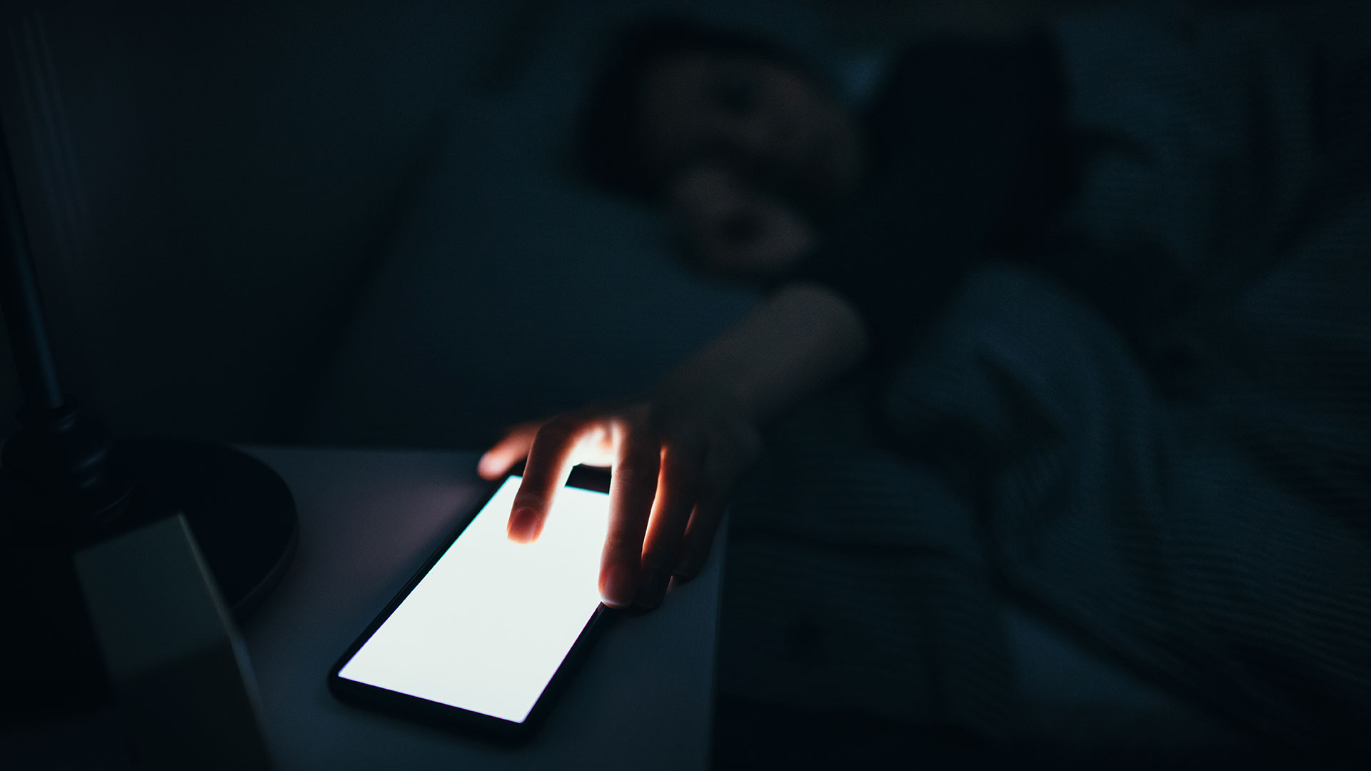 Woman reaching and switching off disturbing calls from work on smartphone while sleeping at midnight