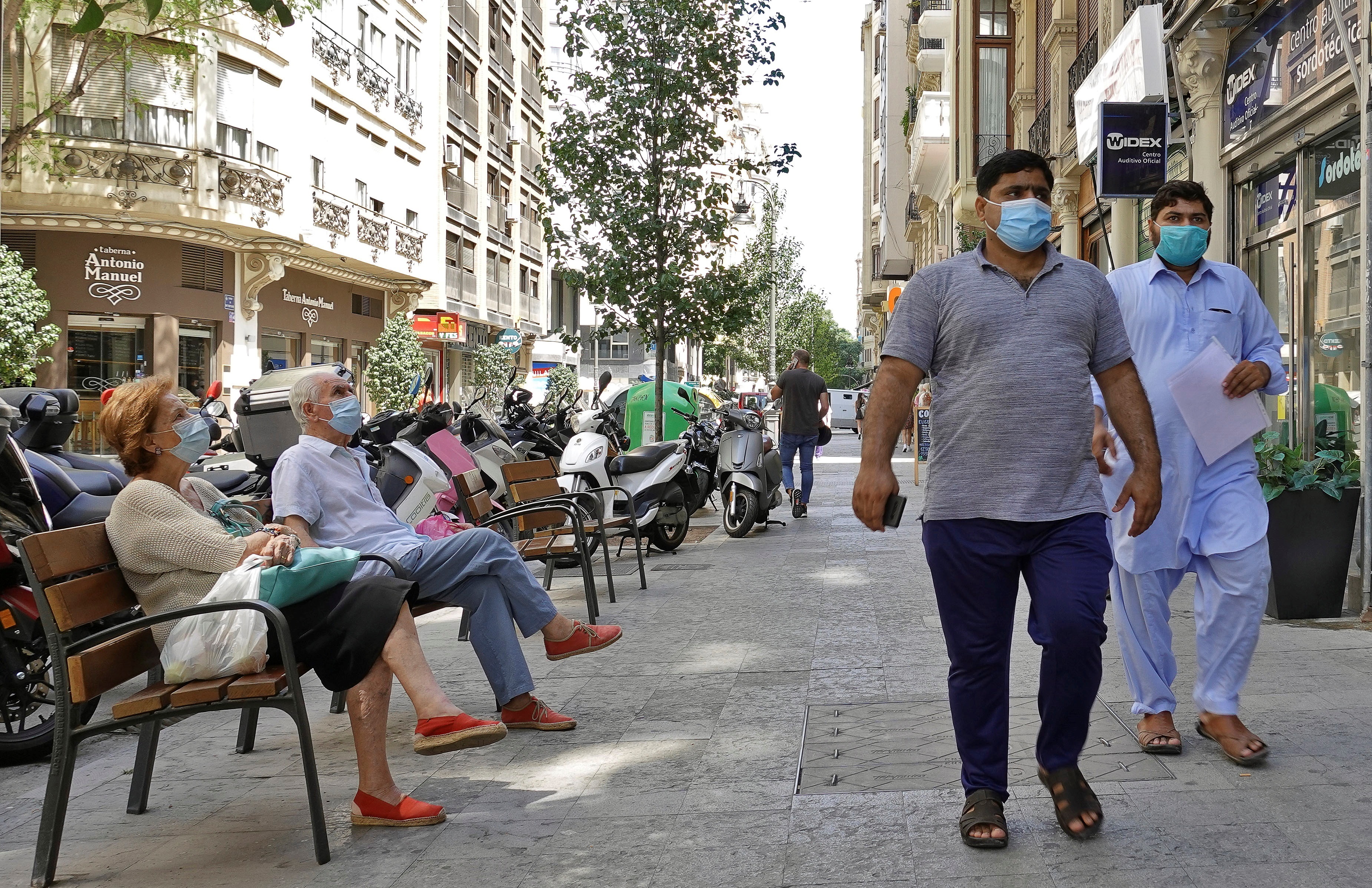 Dos personas descansan sentadas en un banco, mientras otras dos pasan a su lado, todas ellas llevando la obligatoria mascarilla de protección. EFE/Manuel Bruque
