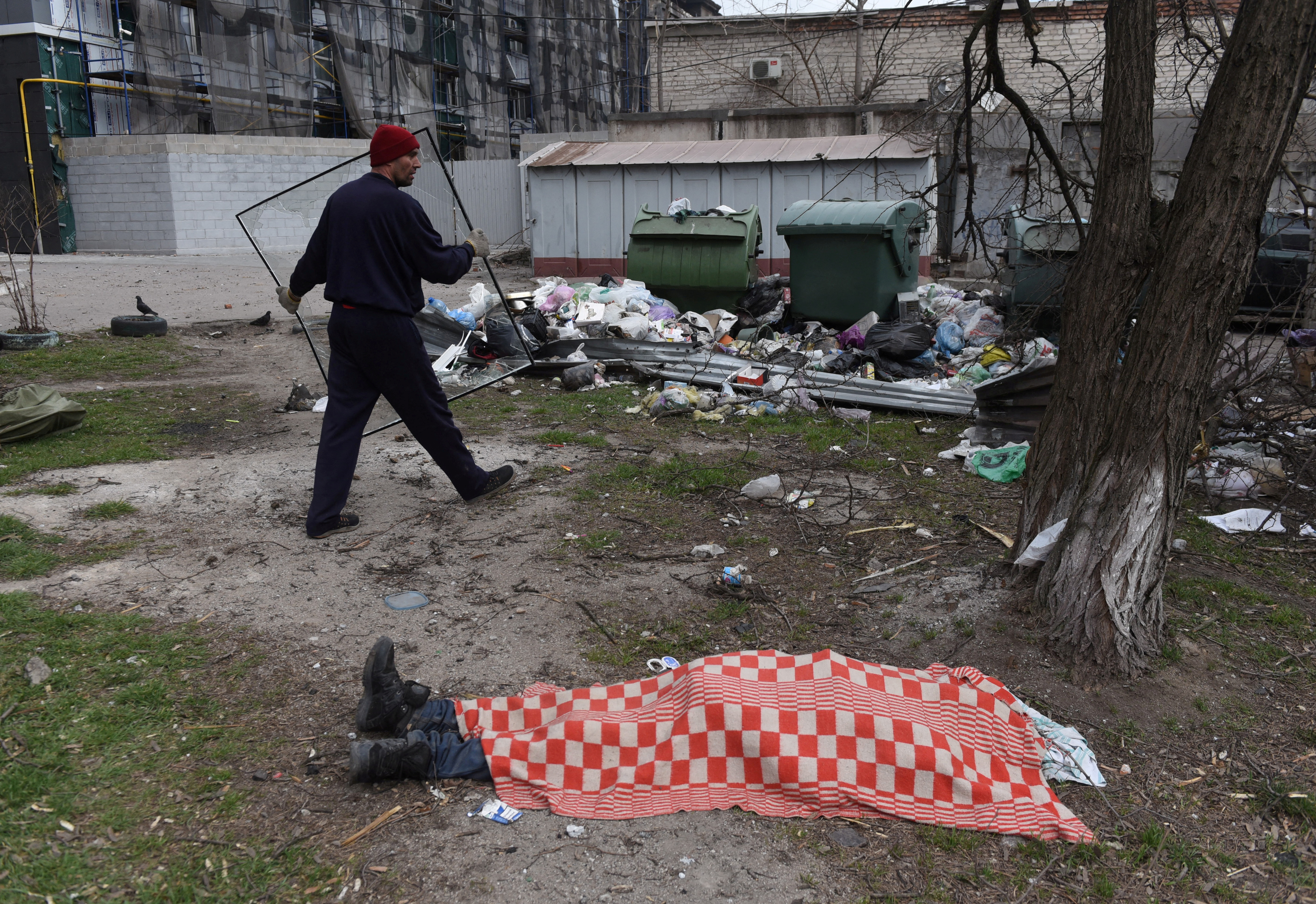 Un residente local lleva una ventana rota junto al cuerpo de una persona asesinada durante el conflicto entre Ucrania y Rusia en una zona residencial en la ciudad portuaria del sur de Mariupol