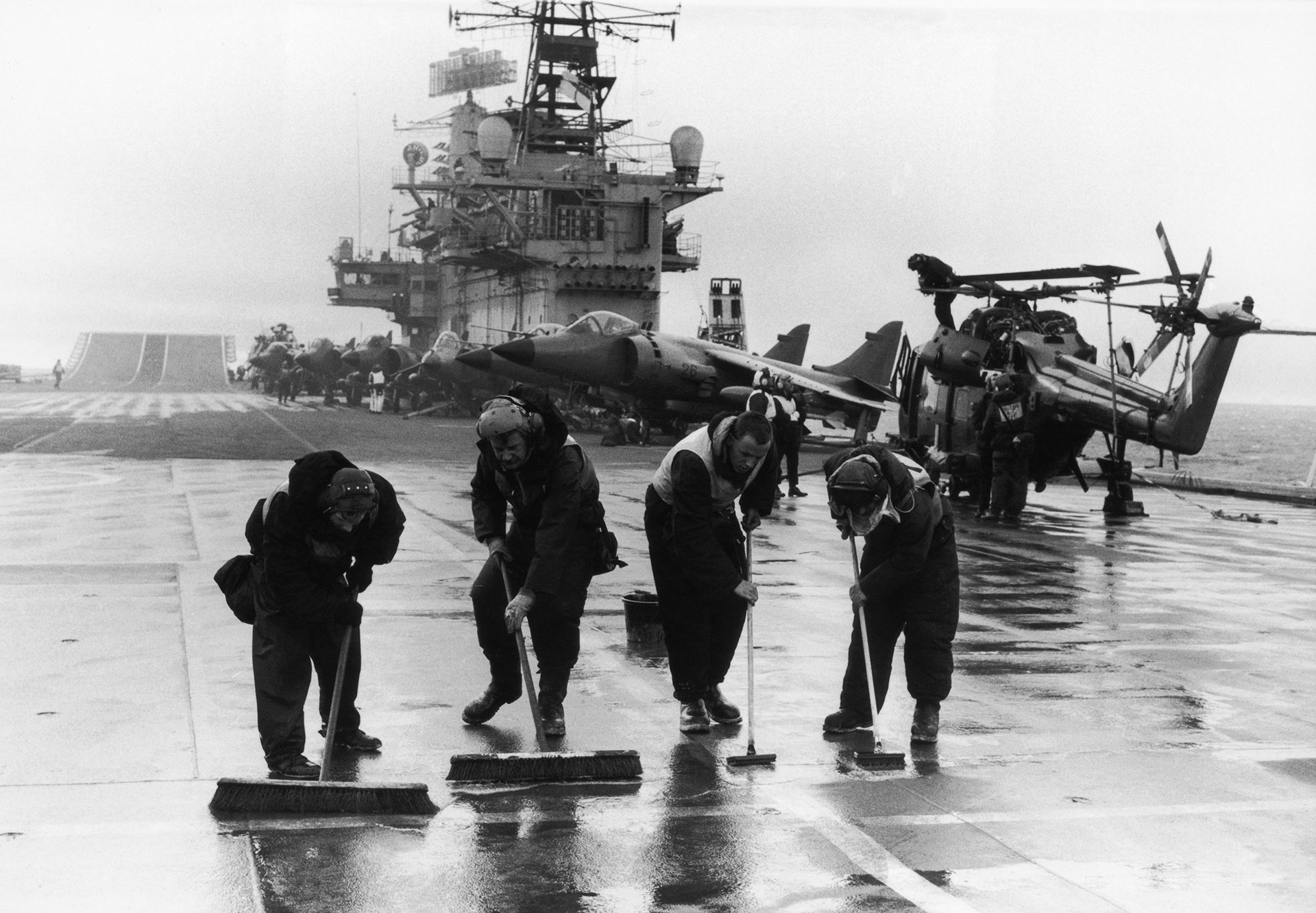 Cuatro hombres limpian la cubierta del portaviones británico HMS Hermes. Llevaba 16 caza navales Sea Harrier, 10 Harrier y 10 helicópteros pesados Sea King, así como un escuadrón de Servicio Aéreo Especial (SAS) y uno de Royal Marines, mayo de 1982 (Martin Cleaver/Pool/Getty Images)