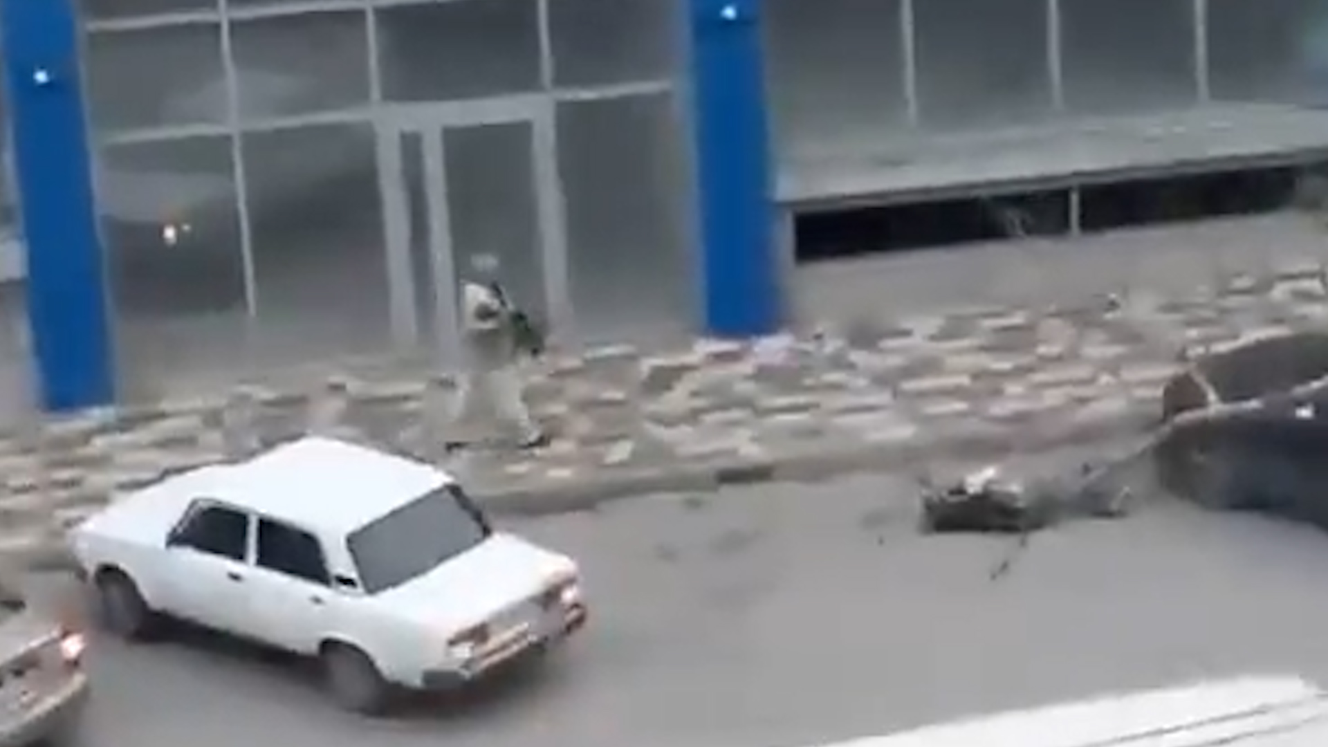 El atacante camninado a las afueras de un centro comercial, portando una ametralladora mientras una víctima yace en el suelo 