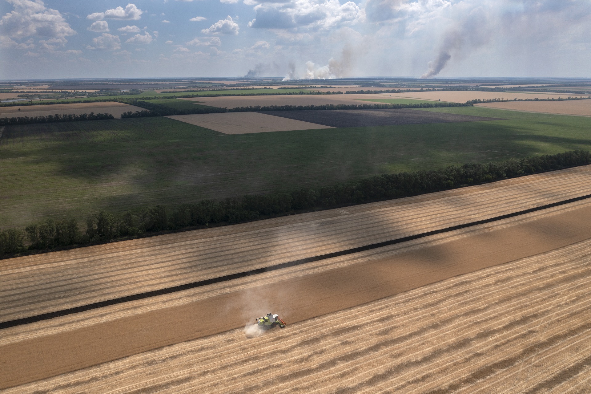 Columnas de humo se alzan al fondo durante fuertes combates, mientras un agricultor cosecha un campo