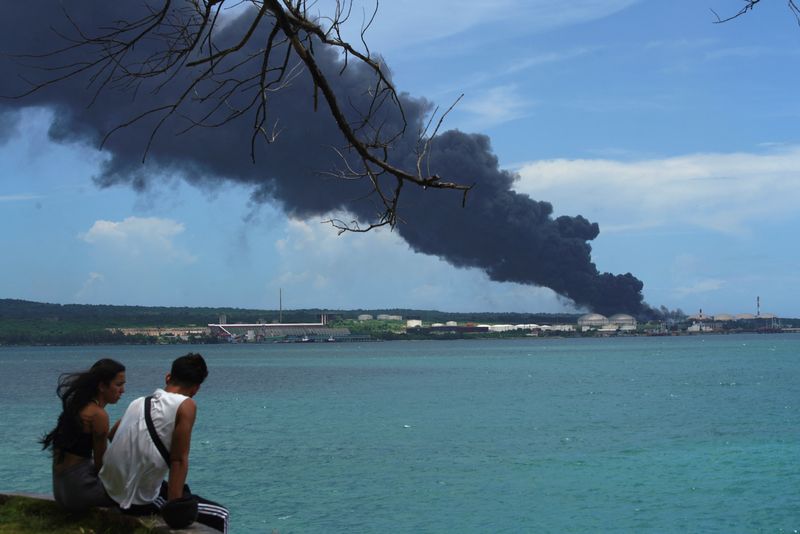 Una pareja se sienta cerca del mar mientras el humo se eleva sobre los tanques, el domingo (Reuters)