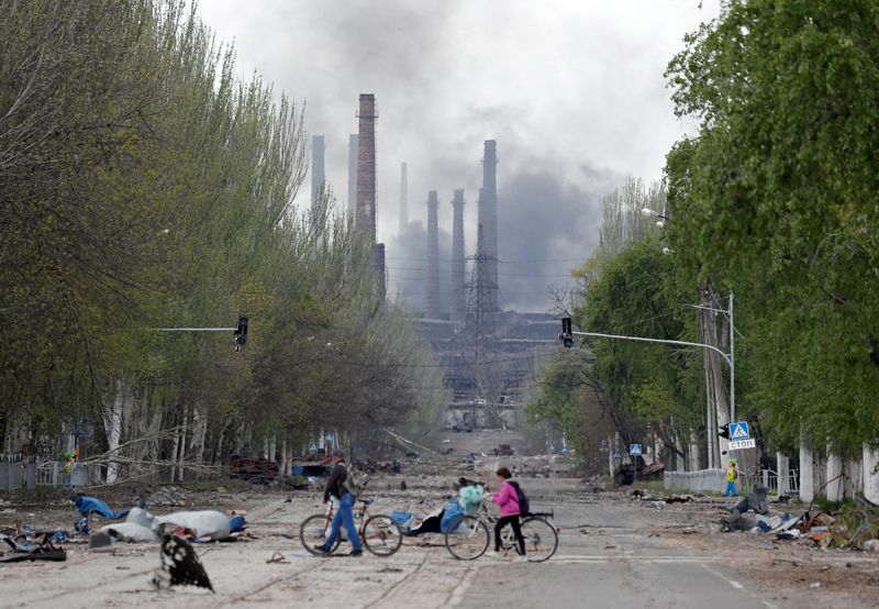 Varias personas cruzan una calle con sus bicicletas con una columna de humo elevándose entre las columnas de la planta siderúrgica Azovstal durante la invasión rusa de Ucrania en Mariúpol, Ucrania, el 2 de mayo de 2022. REUTERS/Alexander Ermochenko