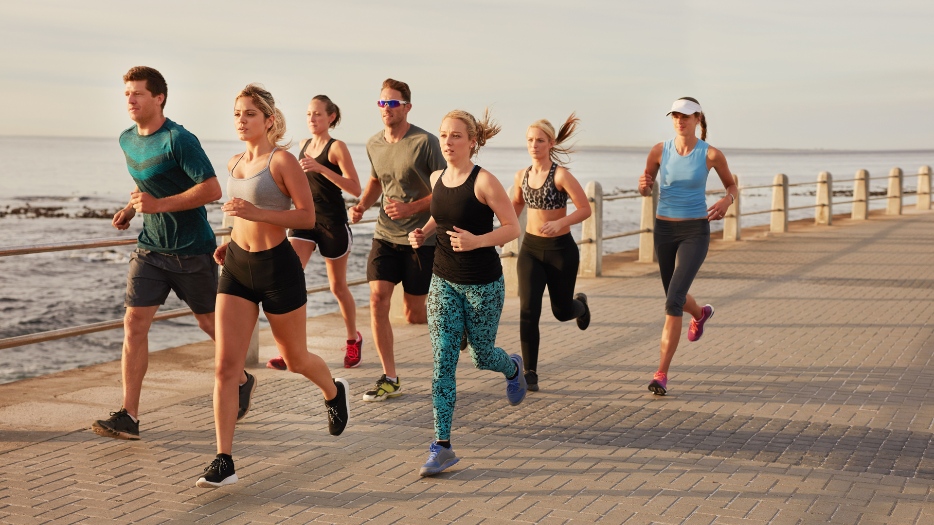 Portrait of young people running along the beach boardwalk by the ocean. Fit young men and women running training outdoors by the seaside.
