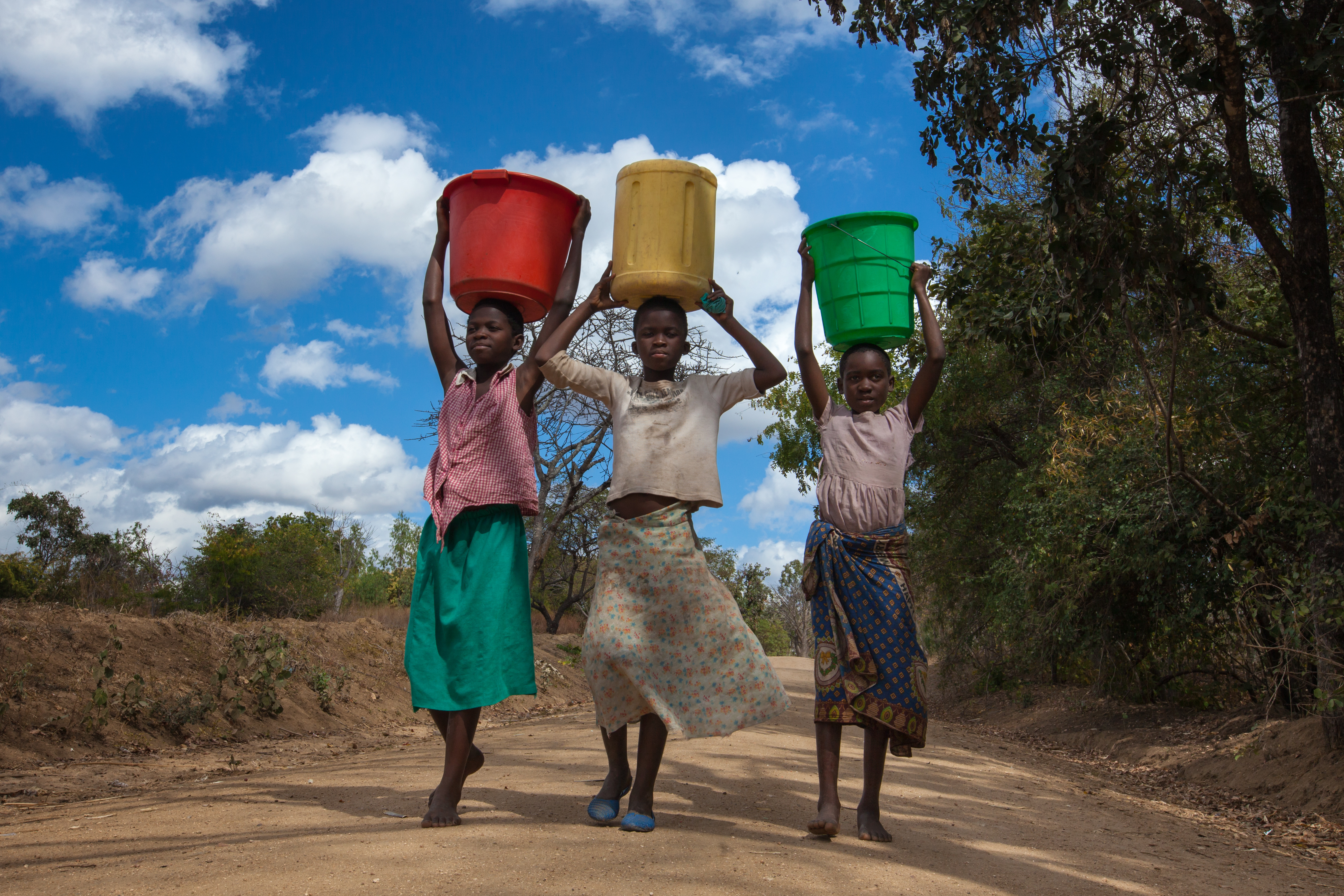 Tres adolescentes cargando cubos de agua de colores en un camino de tierra, regresando de su viaje al pozo más cercano, en el distrito de Balaka, en el sur de Malawi, bajo un cielo azul con nubes