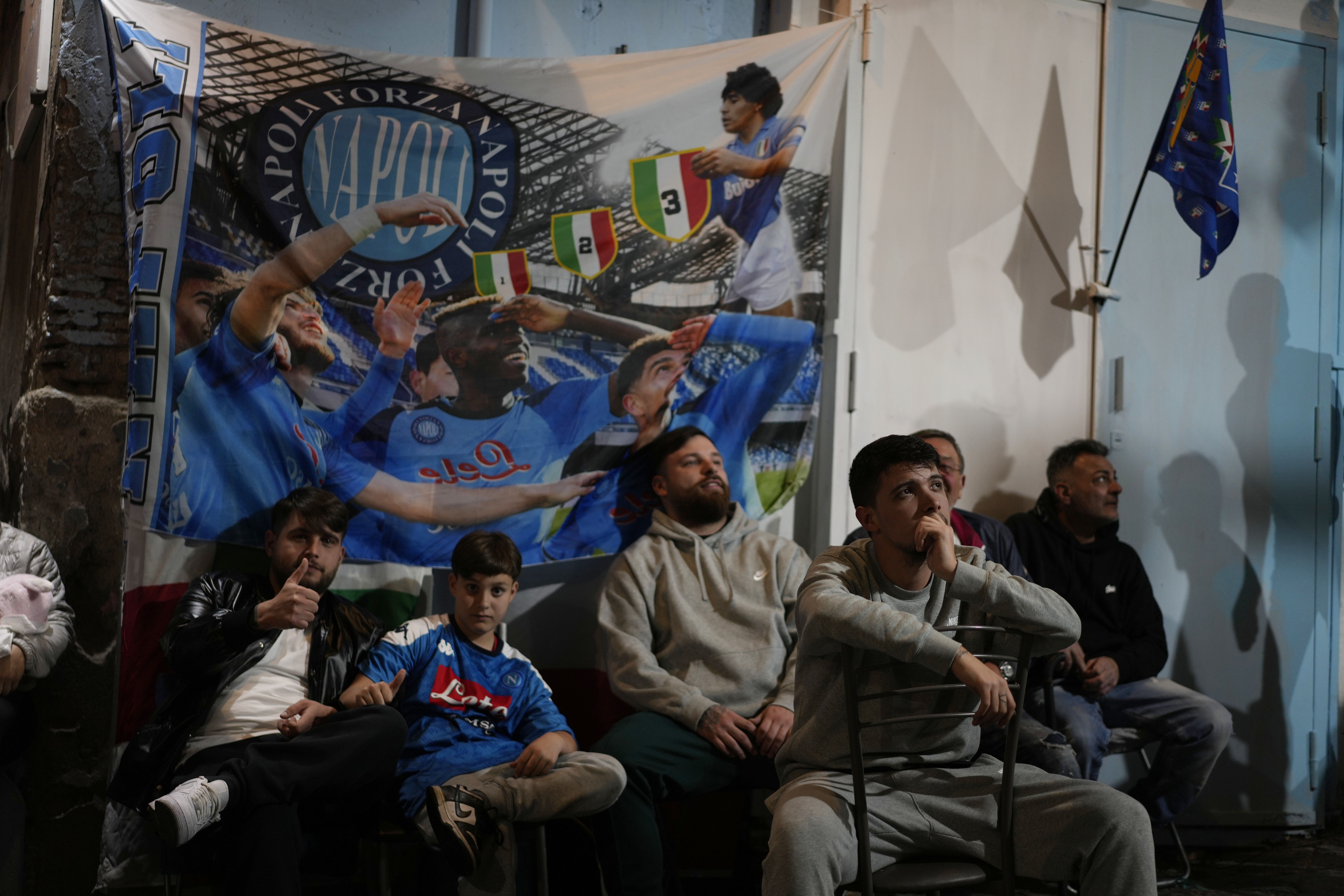 Los hinchas mirando el partido y detrás asoma un dibujo de Diego Armando Maradona