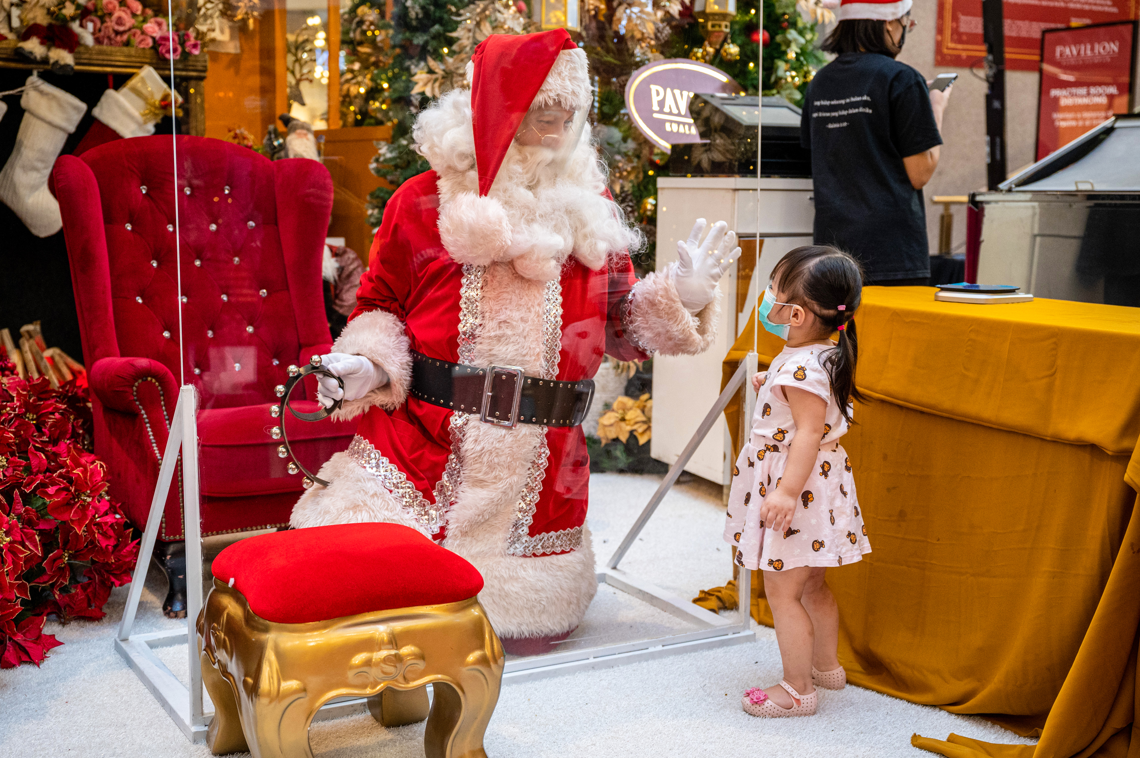 Un hombre vestido con un traje de Papá Noel, usando un protector facial y arrodillado detrás de una barrera transparente, saluda a una niña en un centro comercial de Kuala Lumpur (Foto de Mohd RASFAN / AFP)
