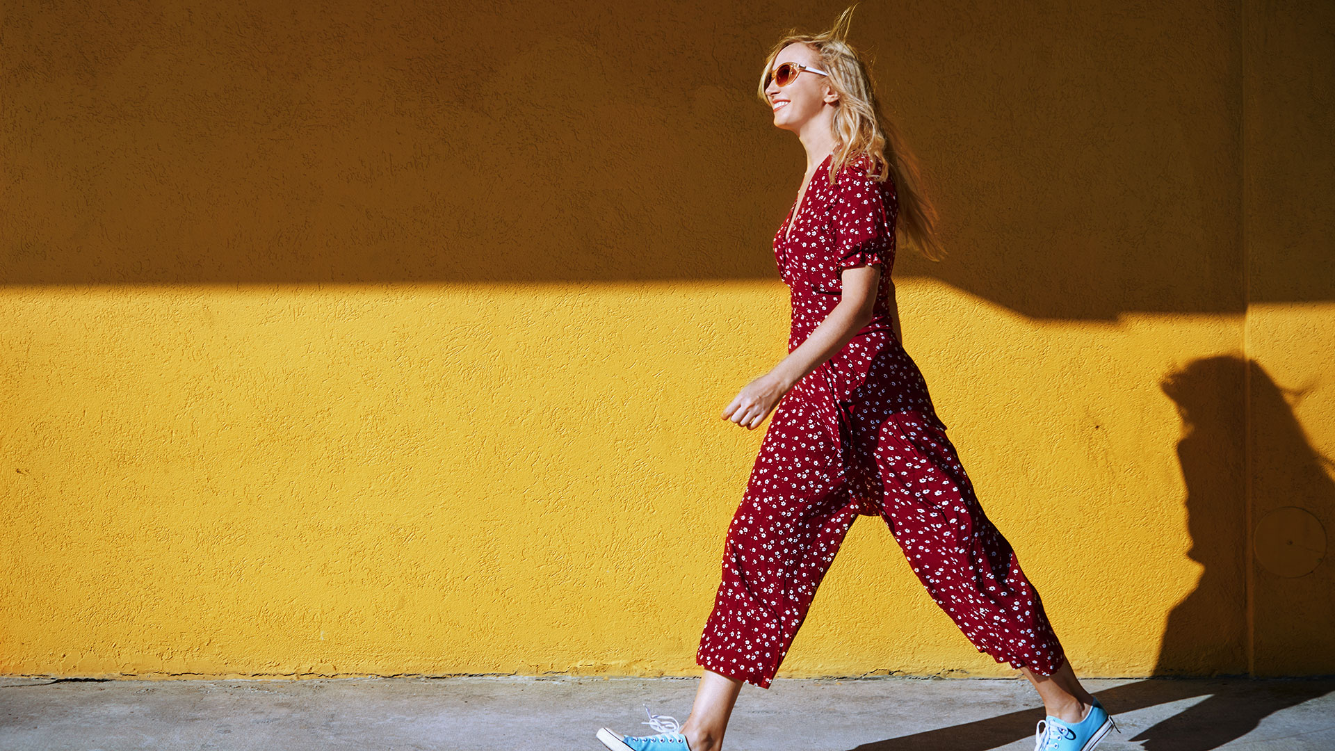 Blond woman wearing burgundy jumpsuit and sunglasses while walking against the yellow wall