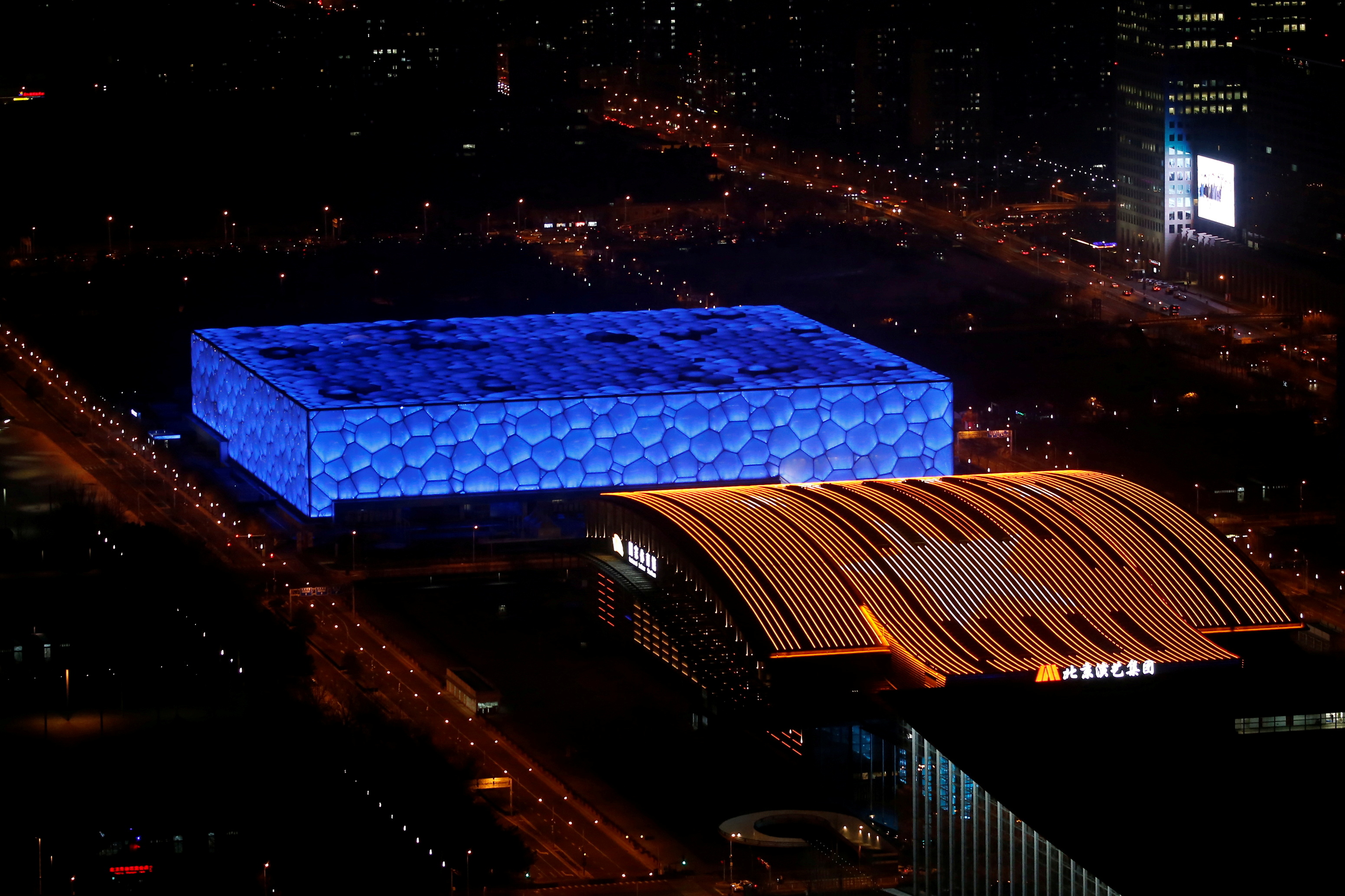 FILE PHOTO: National Aquatics Center, known colloquially as the "Water Cube", is seen next to a building lit up with an image of Chinese national flag, a year ahead of the opening of the the 2022 Winter Olympic Games, in Beijing, China February 4, 2021. Built for the 2008 Summer Olympics, the aquatics center, which will host curling events in the 2022 Winter Olympics, has been given a new nickname "Ice Cube". REUTERS/Tingshu Wang/File Photo