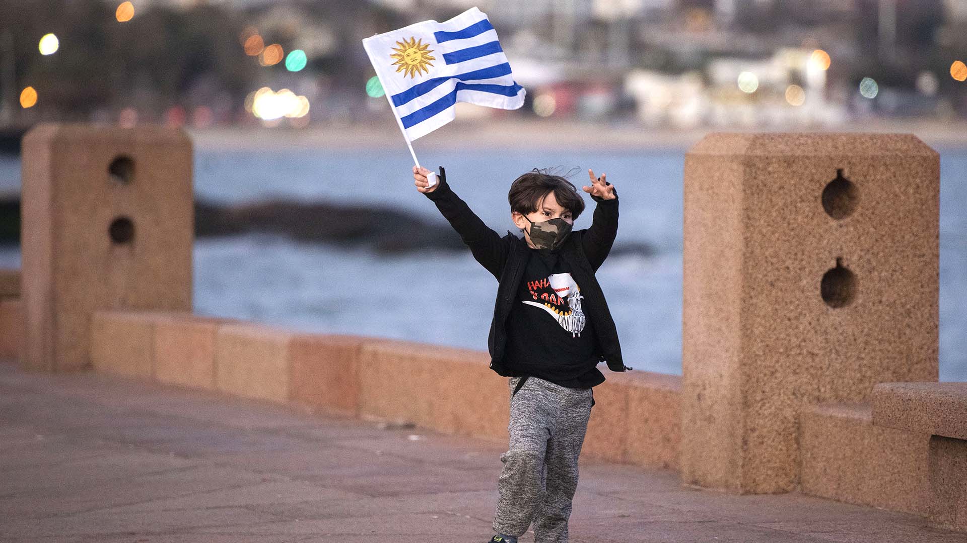 Un niño con una mascarilla protectora ondea una bandera nacional uruguaya mientras juega en el paseo marítimo de Montevideo, Uruguay, el jueves 18 de junio de 2020. (Foto AP / Matilde Campodonico)