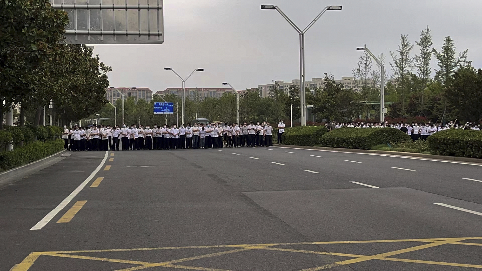 En esta foto publicada por Yang el domingo 10 de julio de 2022, personal de seguridad vestidos de civil se paran en la carretera mientras la gente protesta en la entrada de una sucursal del banco central de China en Zhengzhou. (AP Photo/Yang)