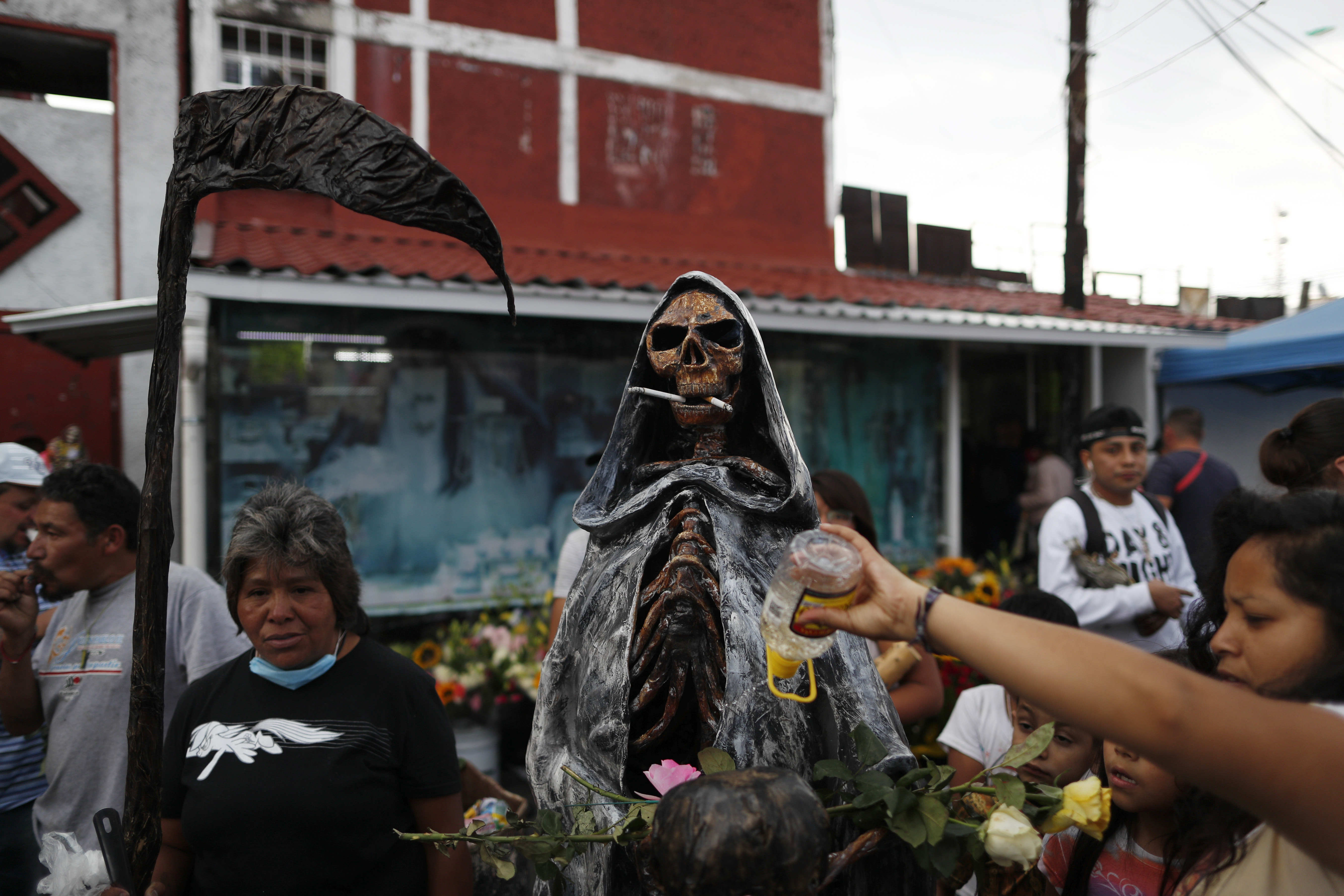 Una mujer arroja alcohol sobre una estatua de la Santa Muerte, o la Santa de la Muerte, cuya boca sostiene dos cigarrillos encendidos que dejaron otros devotos, fuera de su altar en el barrio de Tepito, Ciudad de México, el lunes 1 de junio de 2020. (Foto: AP / Rebecca Blackwell)