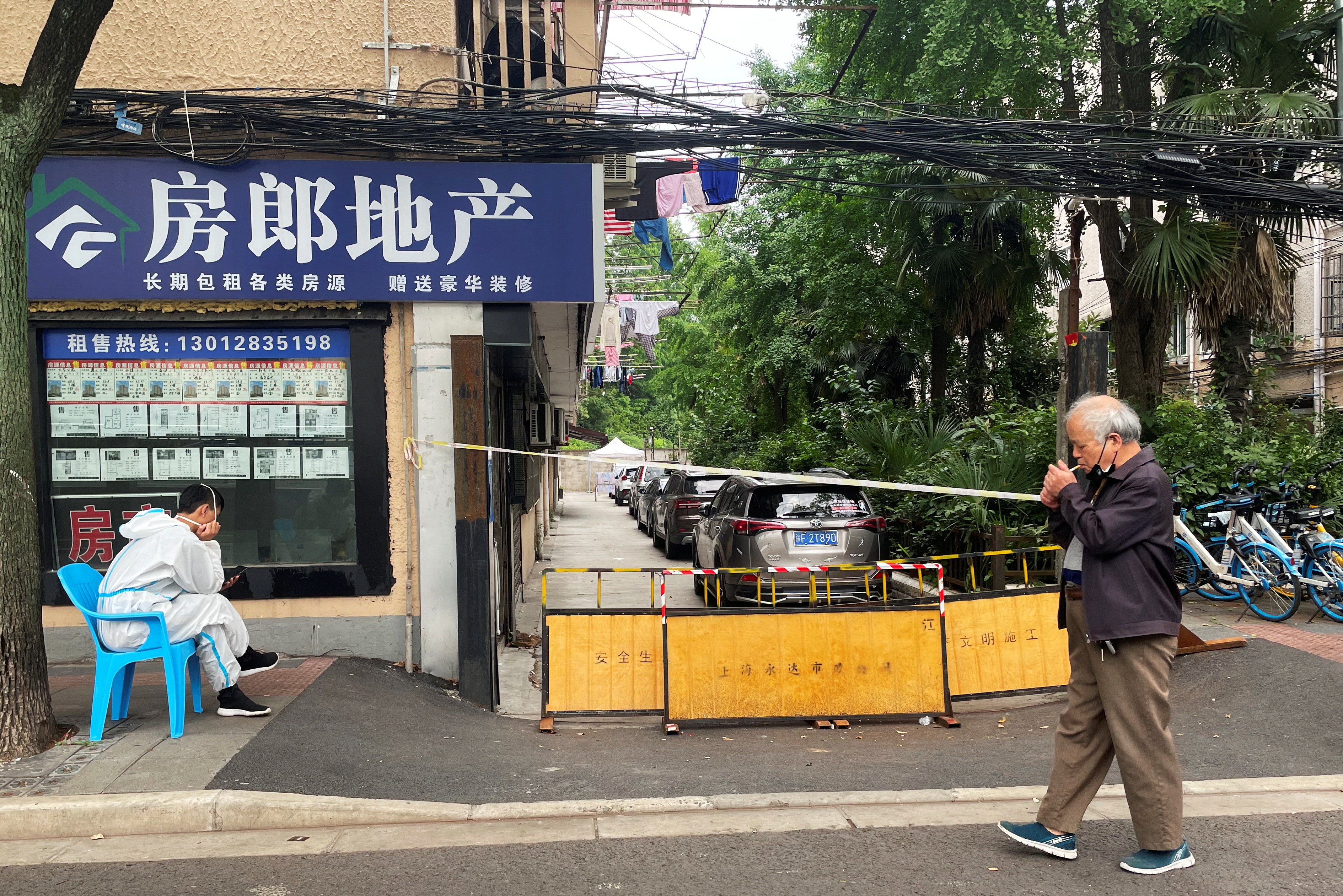 Un trabajador con traje de protección se sienta cerca de una entrada bloqueada a un complejo residencial mientras un hombre pasa por delante, en medio del brote de la enfermedad del coronavirus (COVID-19) en Shanghái (Reuters)