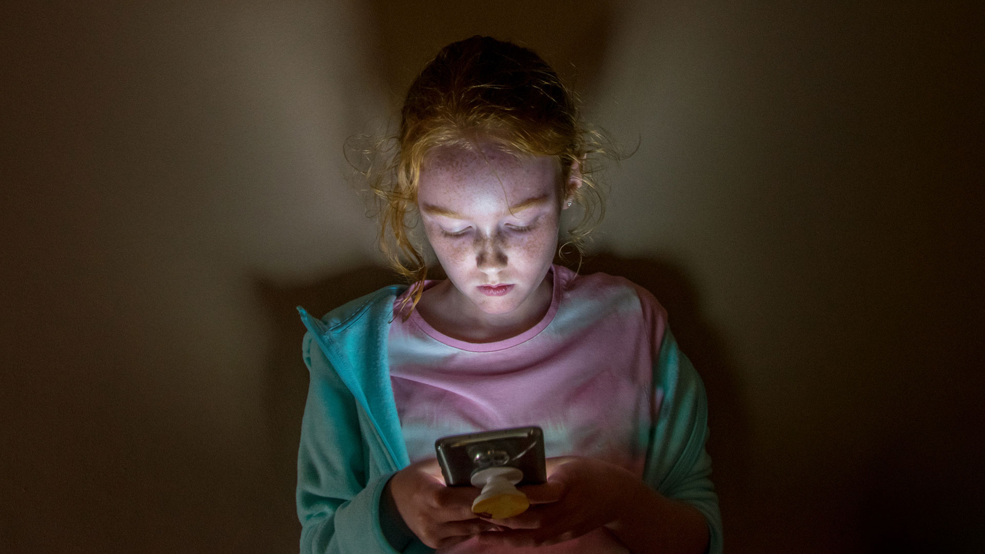 A young long haired redhead girl with face lit up looking into mobile phone screen in a dark room, against a flat wall, with sad facial expression