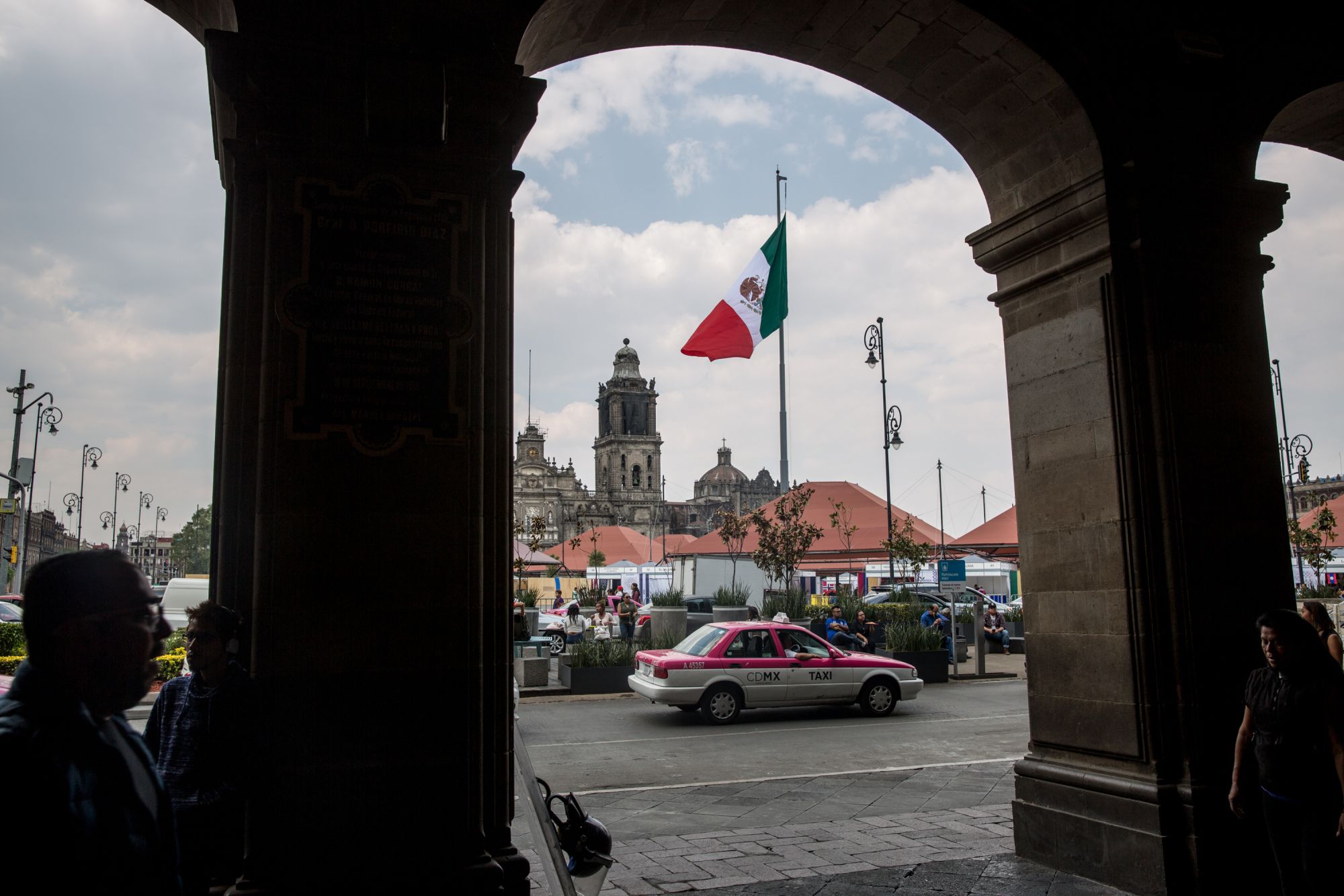 A taxi cab passes in front of the Mexican flag flying at the Plaza de la Constitucion (Zocalo) in Mexico City, Mexico, on Friday, April 13, 2018. Mexico's peso extended losses for a third day amid profit-taking outflows as the currency failed to stay strong past 18.00 key level. Photographer: Alejandro Cegarra/Bloomberg
