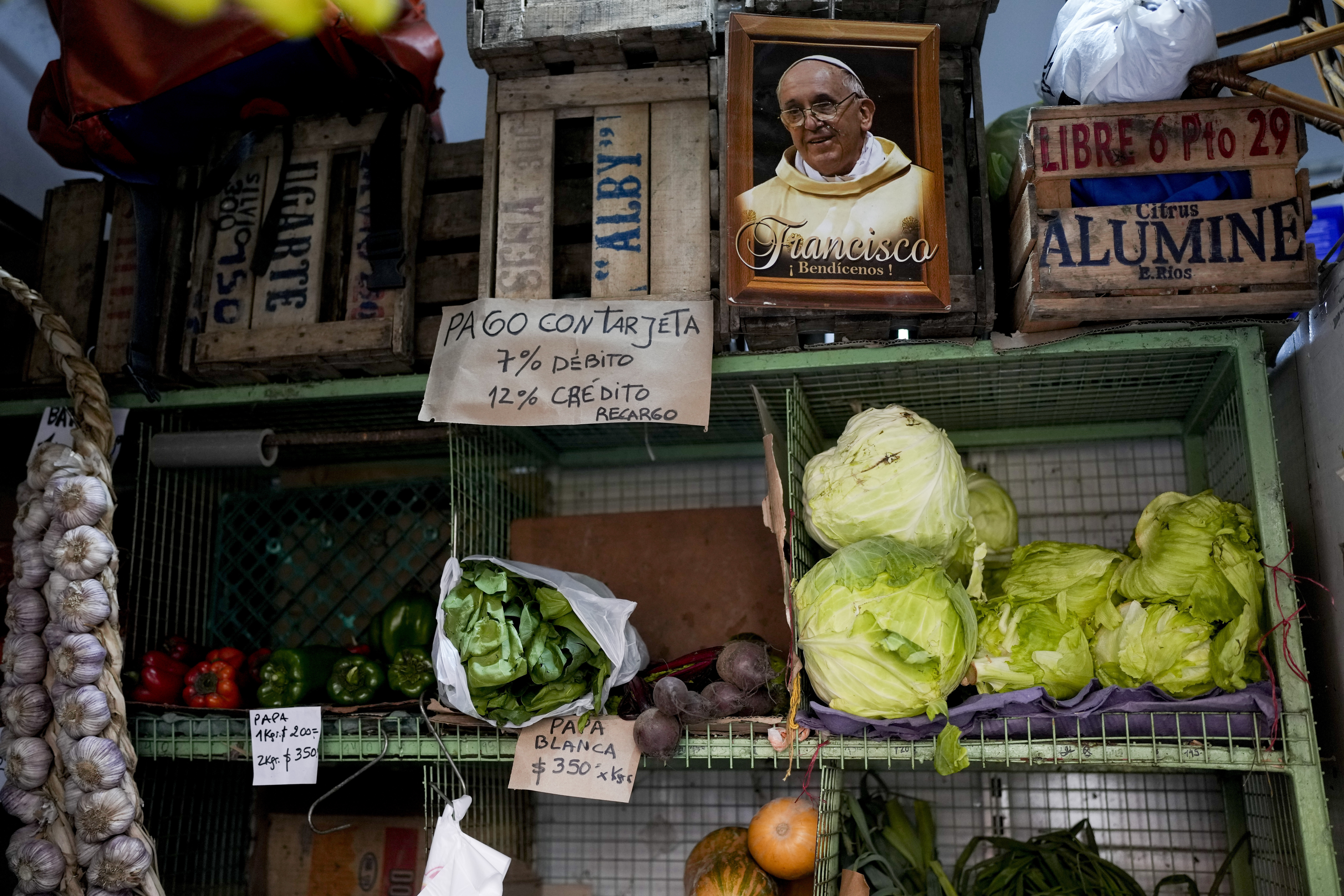 A photograph of Pope Francis hangs next to a sign informing that card payments increase 7% for debit, and 12% for credit, at a fruit and vegetable stall in a market in Buenos Aires, Argentina, May 11, 2023. According to a recent report on food security from the World Bank, the price of food in Argentina suffers year-on-year inflation of 107%.  (AP Photo/Natacha Pisarenko)