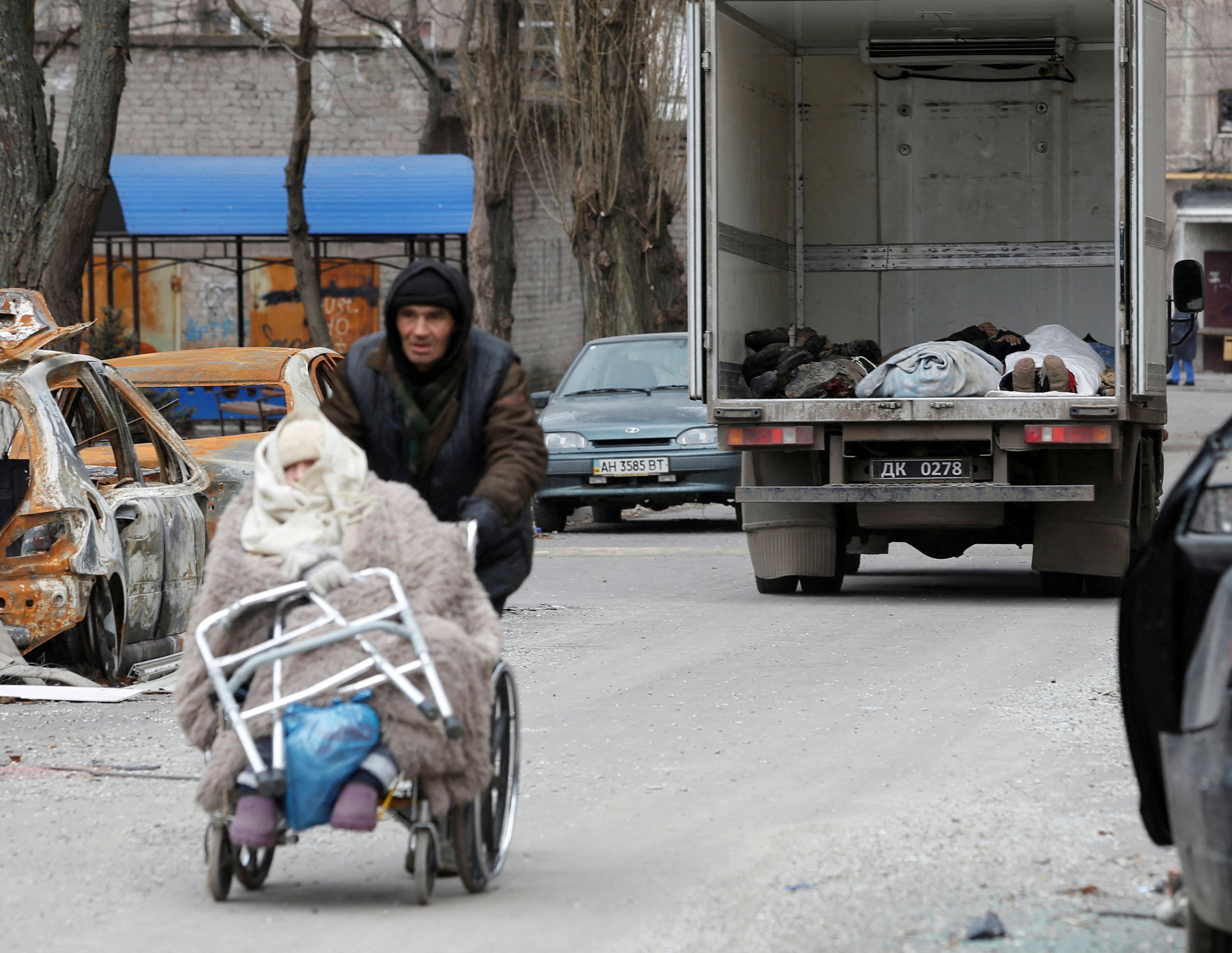 Un hombre empuja a una anciana en silla de ruedas, mientras los cuerpos de los muertos recogidos por miembros del servicio de las tropas prorrusas se ven dentro de un vehículo antes de ser transportados a la morgue durante el conflicto entre Ucrania y Rusia en la ciudad portuaria del sur de Mariupol, Ucrania 4 de abril de 2022. REUTERS/Alexander Ermochenko