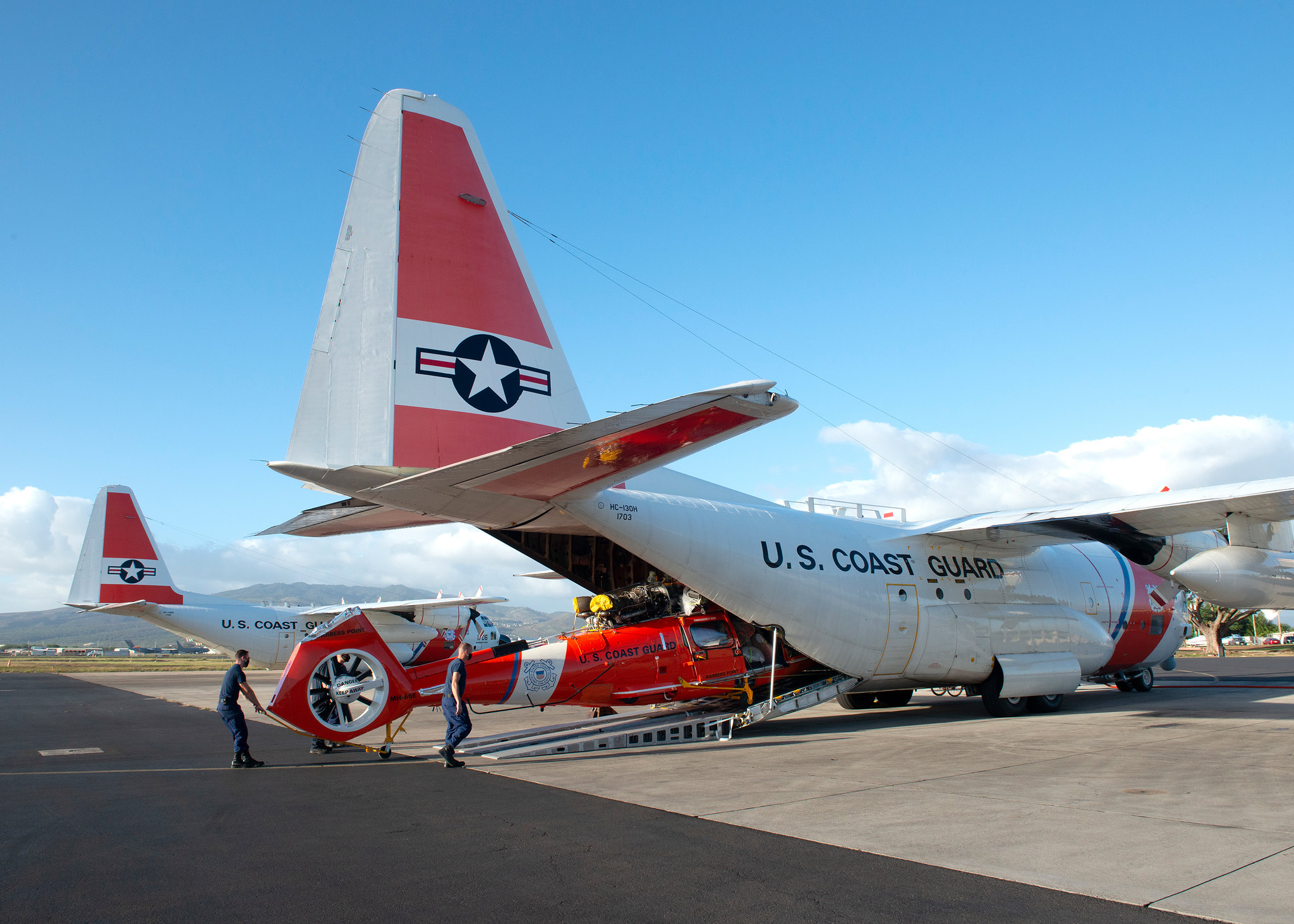 A Coast Guard Air Station Barbers Point aircrew unloads a MH-65E Dolphin helicopter from a HC-130 Hercules airplane on Oahu, Hawaii, Jan. 28, 2021. The helicopter is a new “echo” model with improvements ranging from enhanced digital weather/surface radar to modernization of the avionics from the previous “delta” model. (Petty Officer 2nd Class Matthew West/Coast Guard)