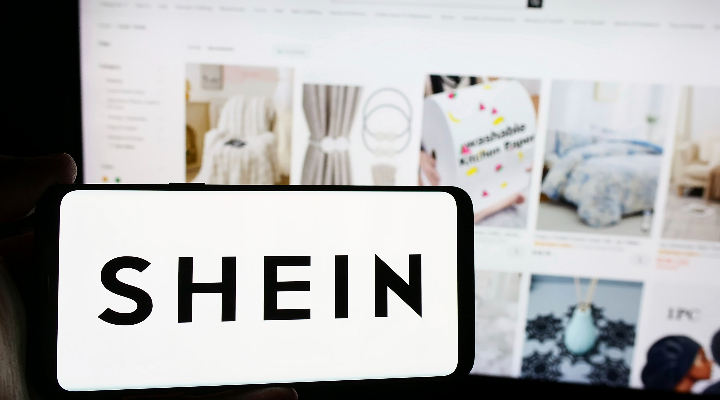 In this photo illustration, a person holds a mobile phone with the logo of fashion e-commerce company Shein in front of business webpage.