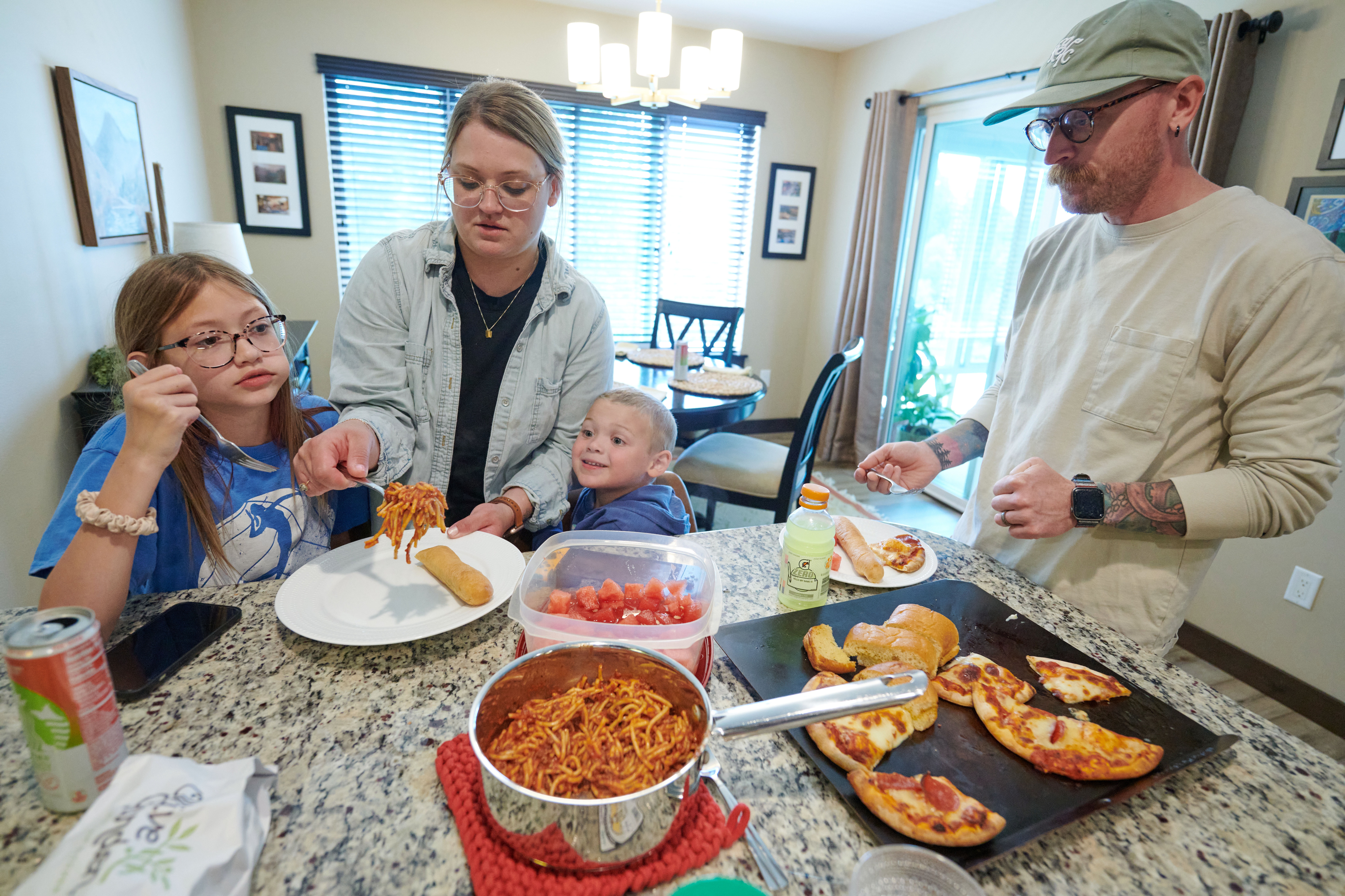 Amber and Neil Petersen serve pasta, pizza and fruit to their children, 11-year-old Eden and 4-year-old Jack, in Iowa City, Iowa. They are standing around a cluttered countertop.