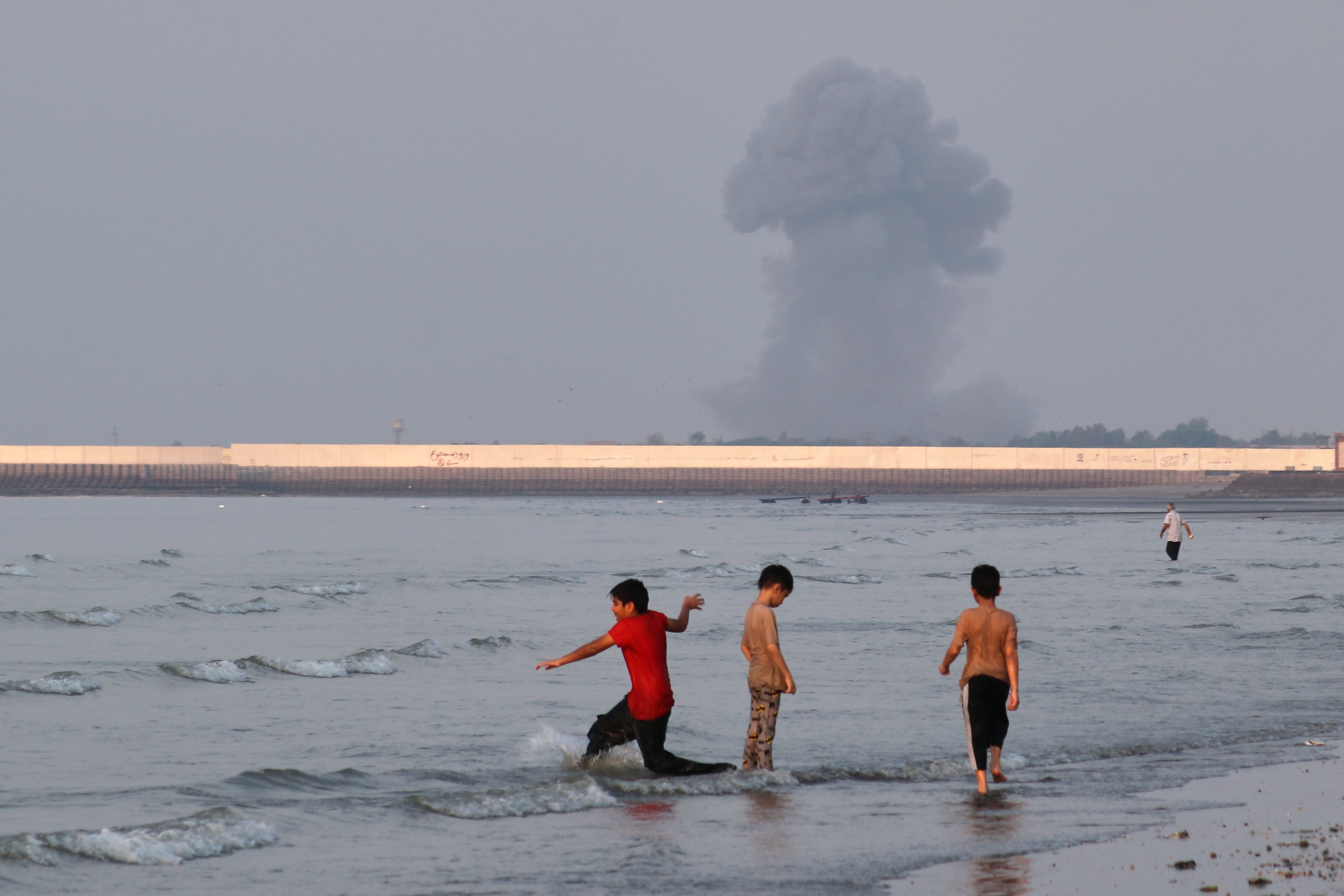Three boys play in the shallow waters of the Strait of Hormuz, as a plume of smoke rises from an explosion in the background, off Bandar Abbas, Iran, Monday, July 13, 2026.