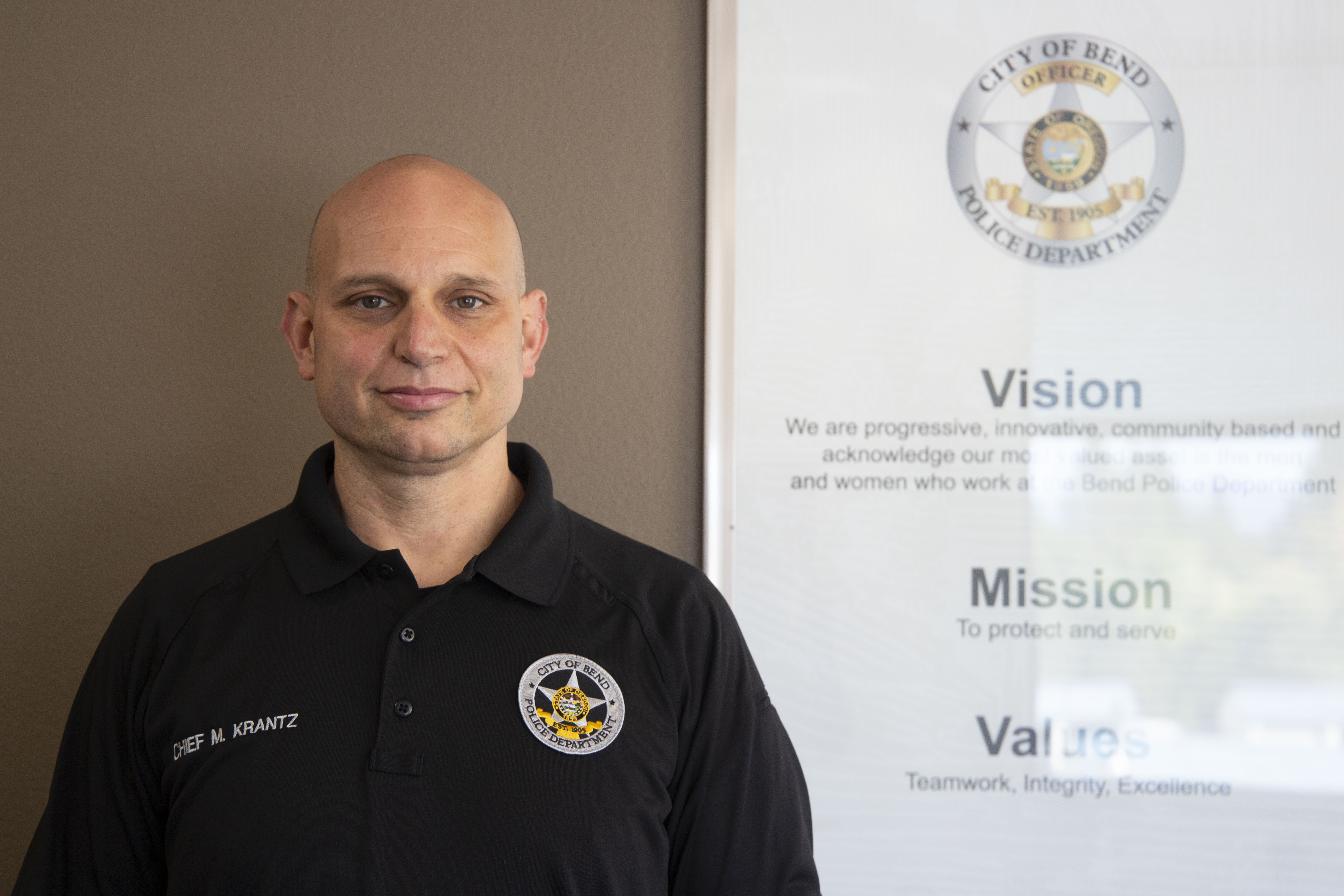A man wears a black polo shirt with the logo of the Bend Police Department on one side of his chest and the words "Chief M. Krantz" on the other.