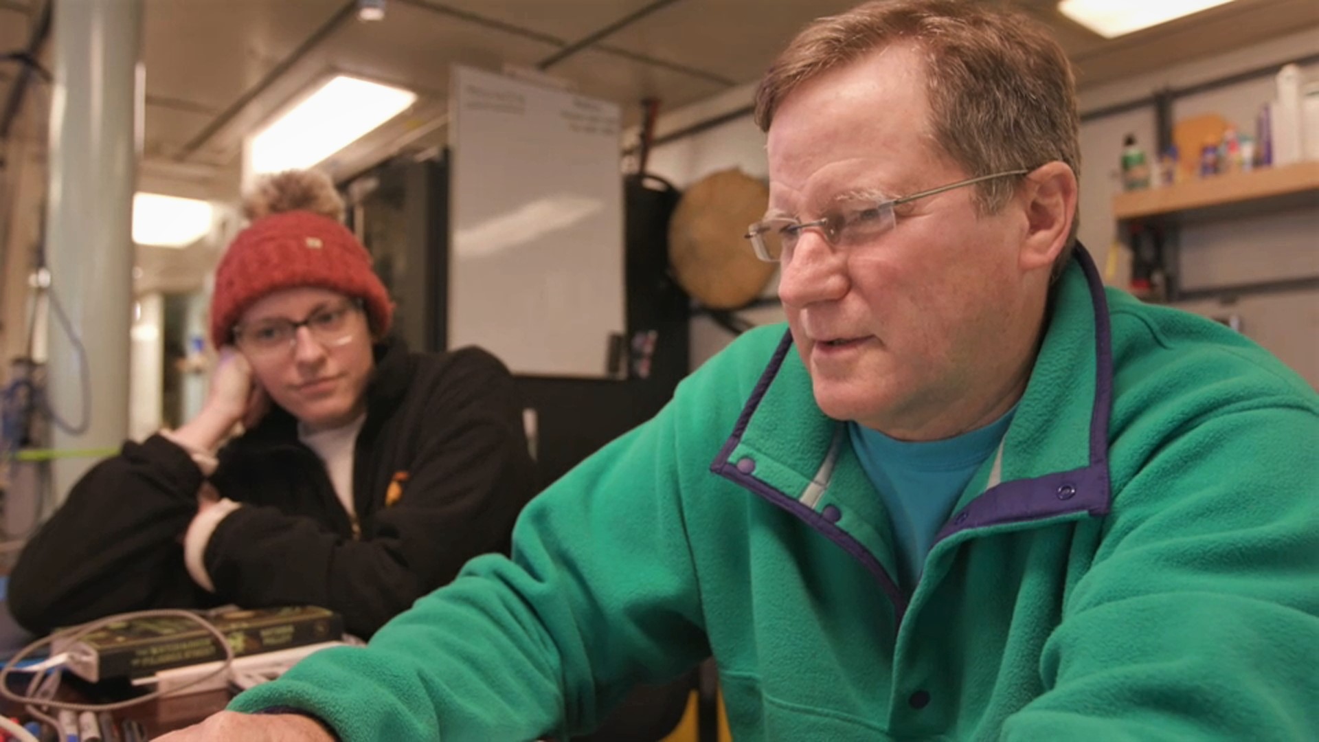 a man with glasses and brown hair in a green sweater and a woman in a red beanie and glasses in a lab