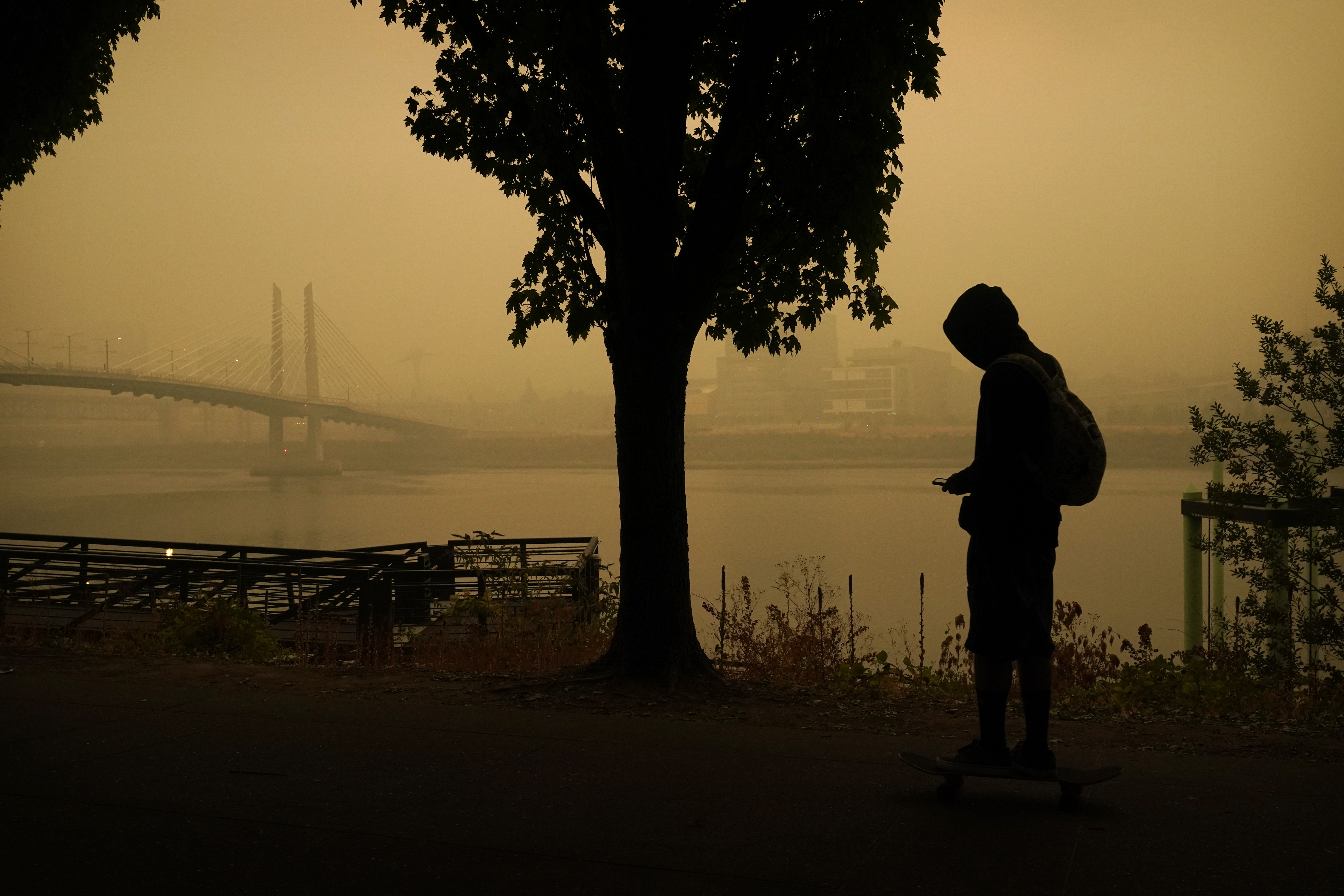 A person rides a skateboard along the Willamette River as smoke from wildfires partially obscures the Tilikum Crossing Bridge, Saturday, Sept. 12, 2020, in Portland, Ore.