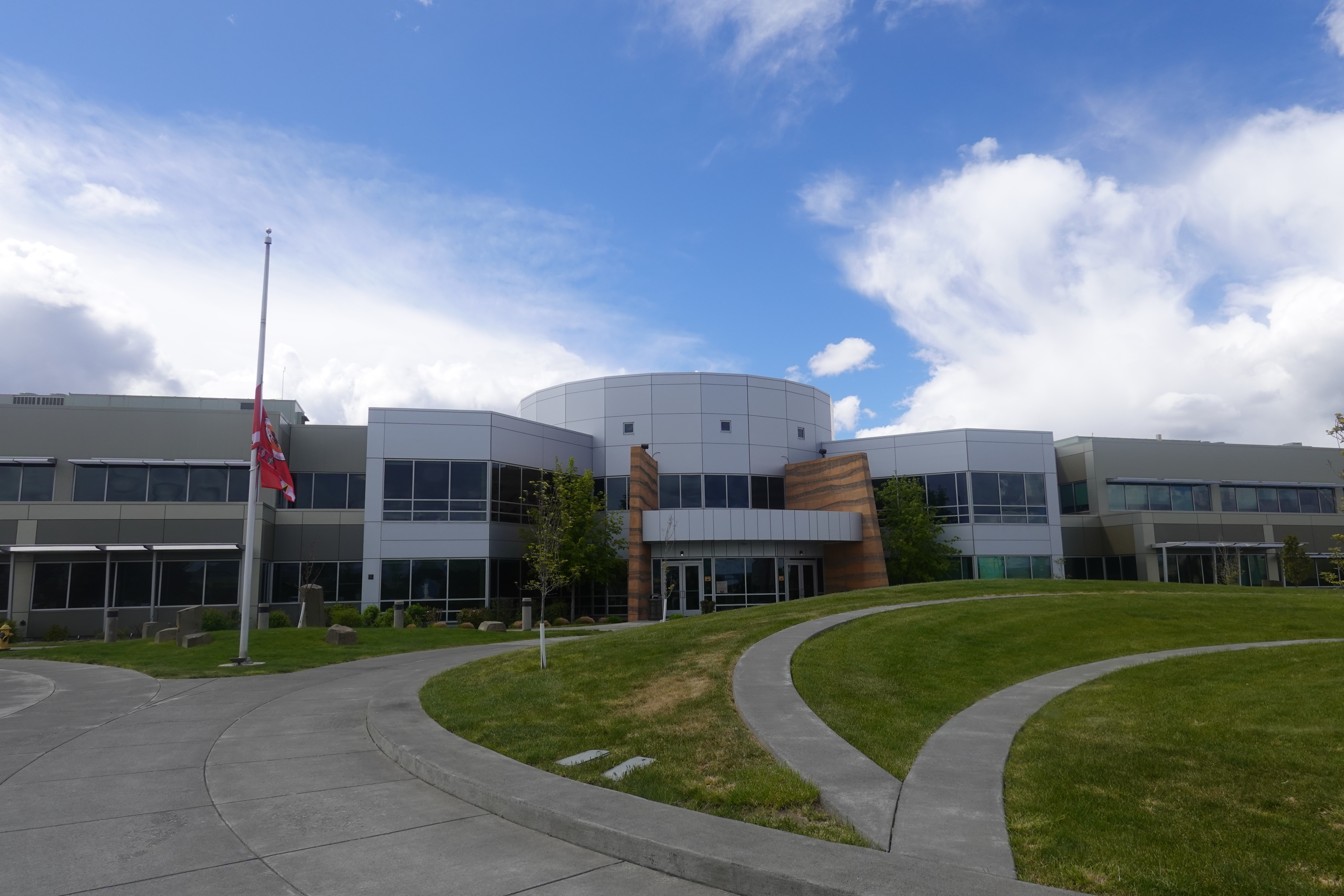 A gray two-story building sits under a cloudy sky as the red flag for the Confederated Tribes of the Umatilla Indian Reservation sits at half-mast.