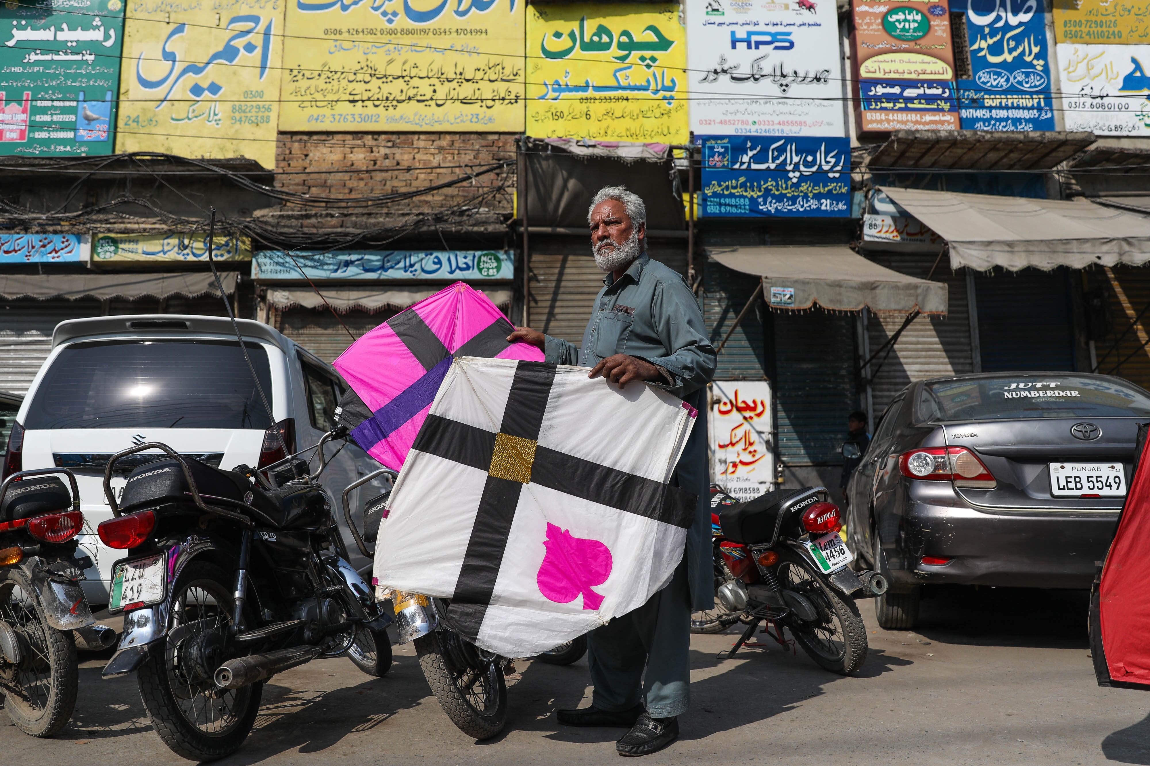 An older man with white hair and a white beard holds a pink kite in one hand and a white one in the other outside a market area near Mochi Gate during Basant.