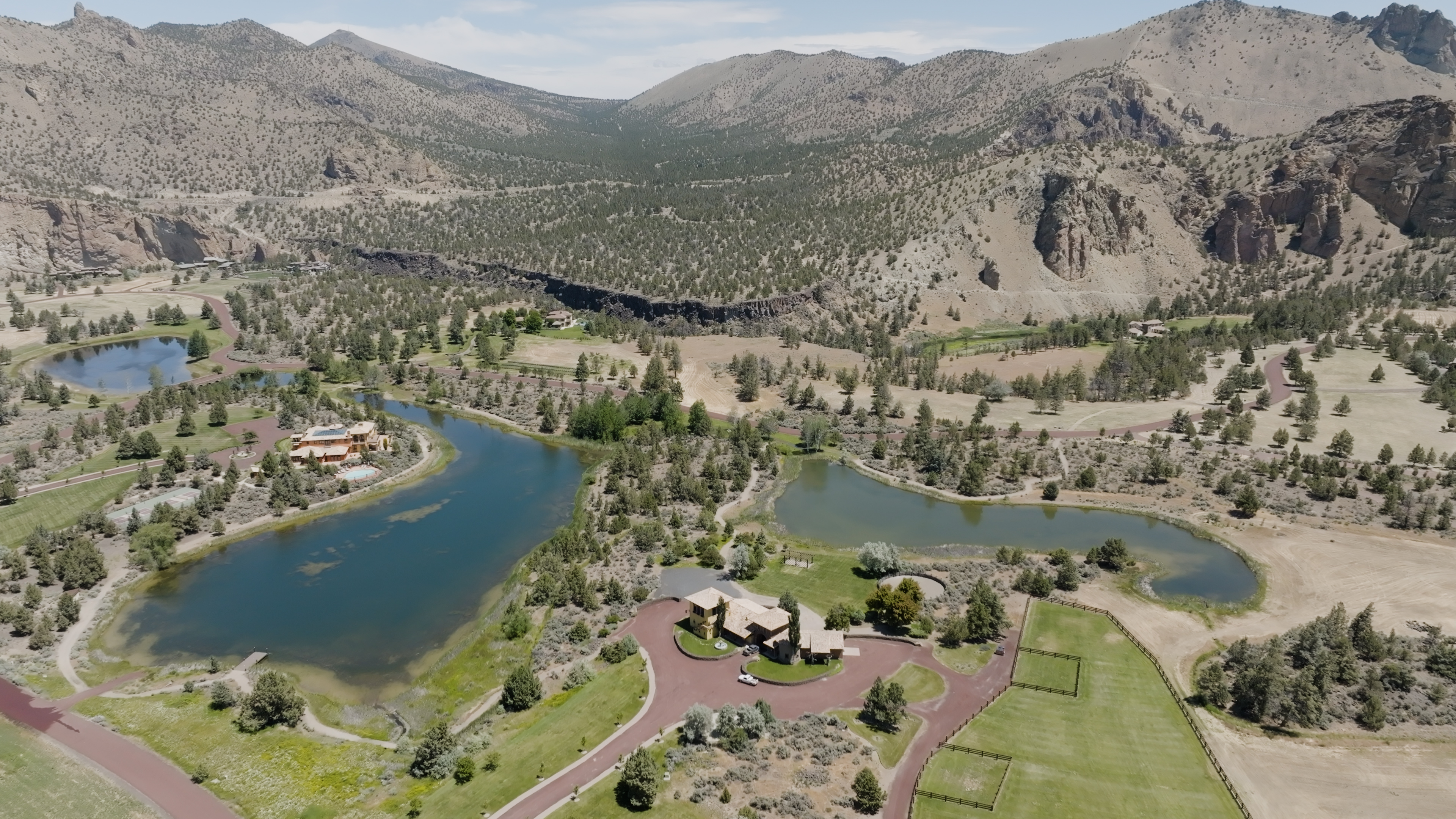 A wide, sweeping landscape photograph showing mansions, large, lush green lawns, pools of water, and mountains in the background.