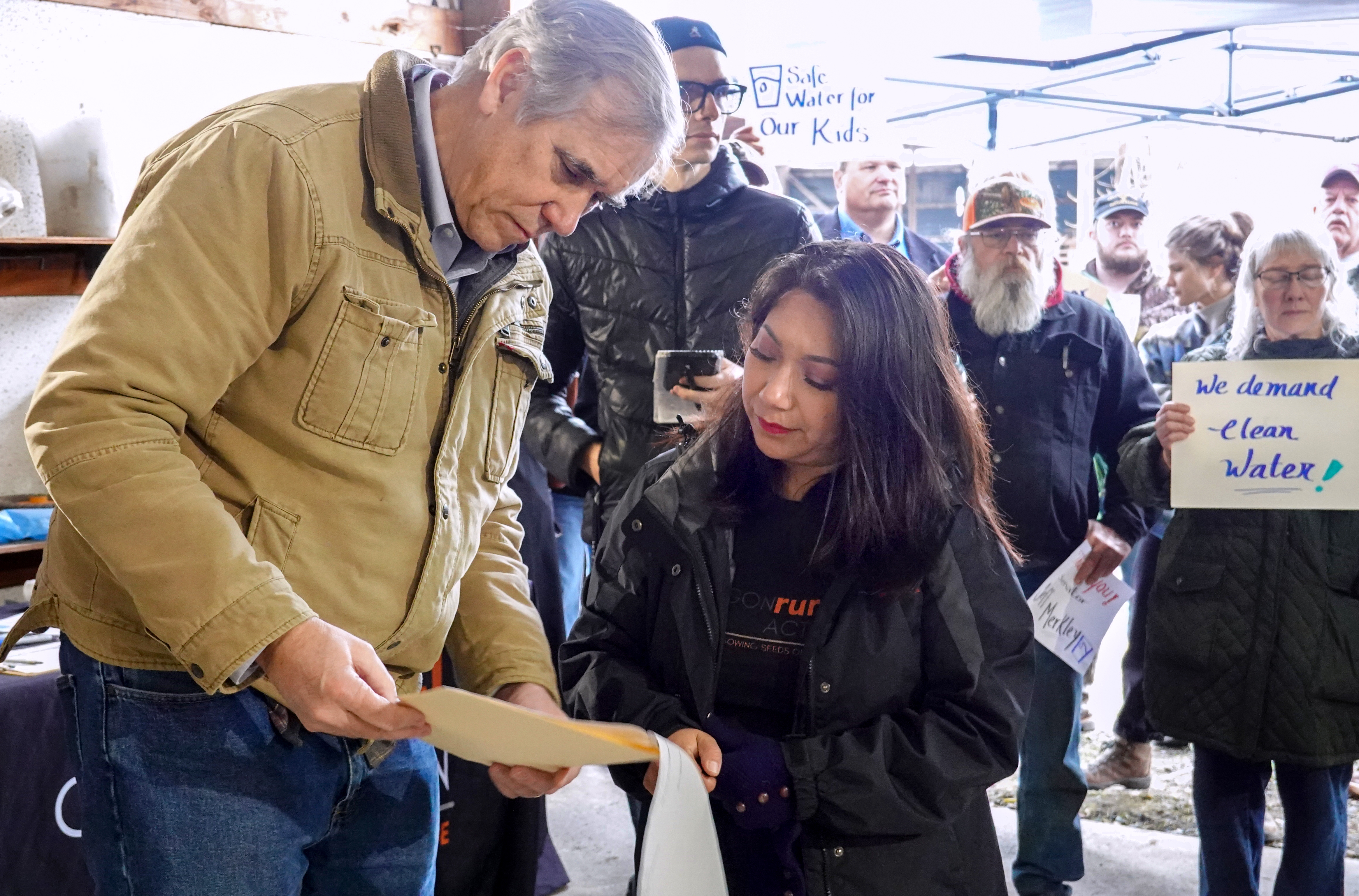Jeff Merkley looks down at some pieces of paper while a woman stands next to him in front of a small crowd of people.