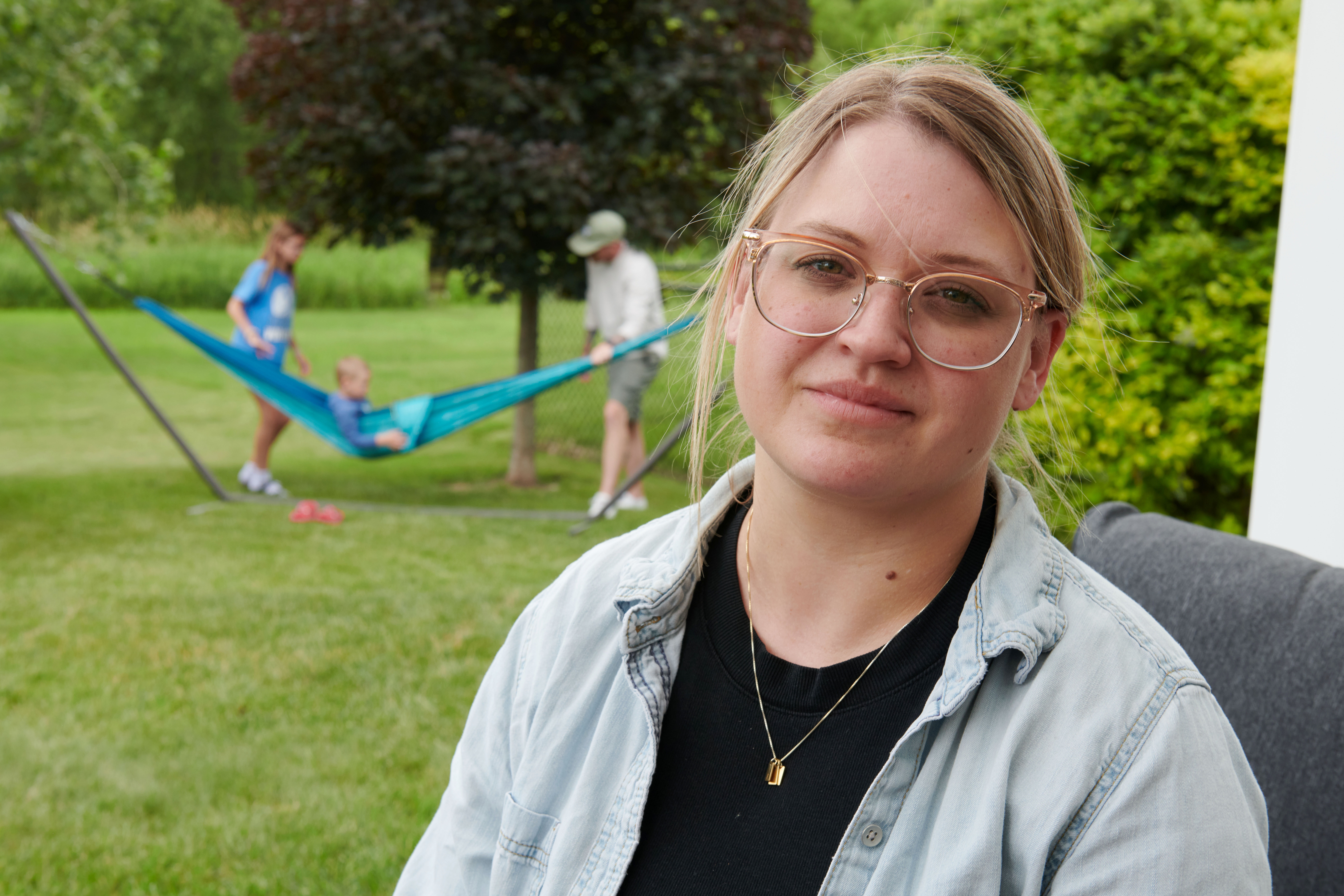 Amber Petersen sits in the backyard of her parents’ home in Iowa City, Iowa. She has blonde hair pulled back and is wearing glasses. Her husband and children are playing in a hammock behind her.