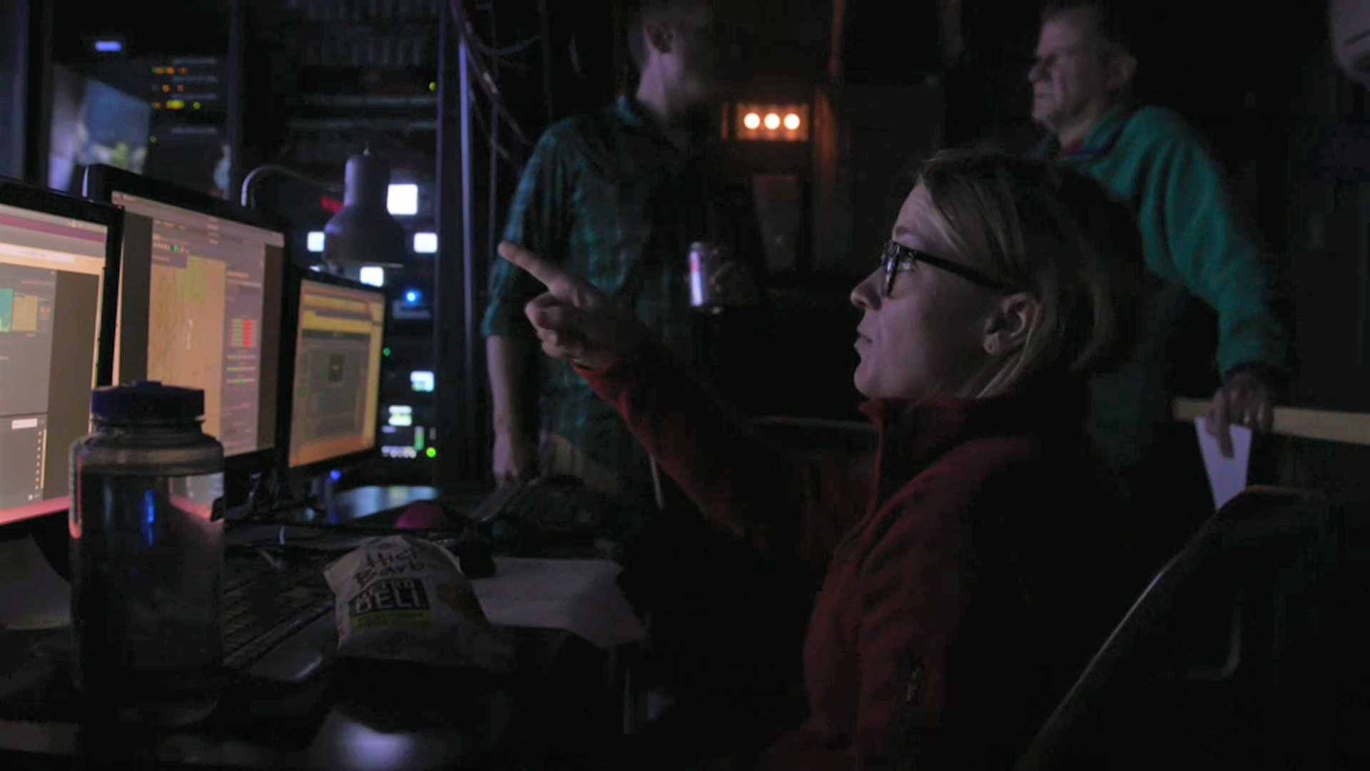 in a dark room with a lot of laboratory equipment and machinery, a woman points at a computer collecting data. 