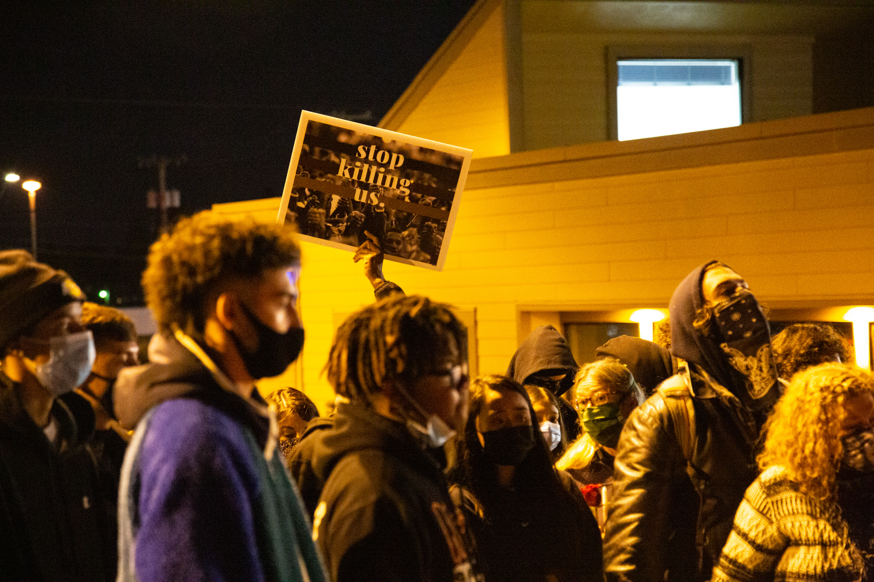 A crowd of people gathers at night wearing masks on their faces. One person holds a sign above the crowd's heads that reads "Stop killing us."